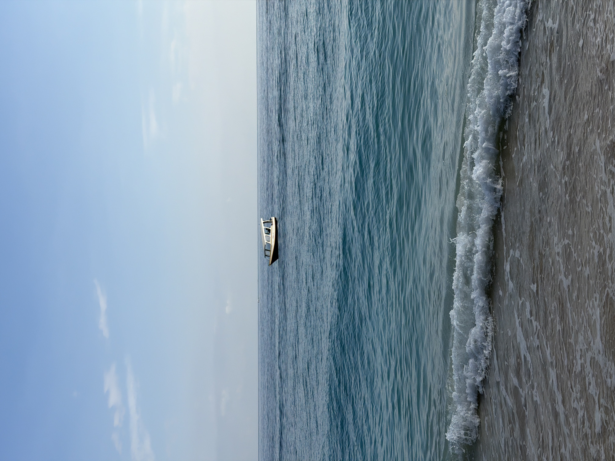 A white and brown motorboat is on a blue ocean under a clear sky with a few white clouds. Waves are breaking on a sandy beach in the foreground.