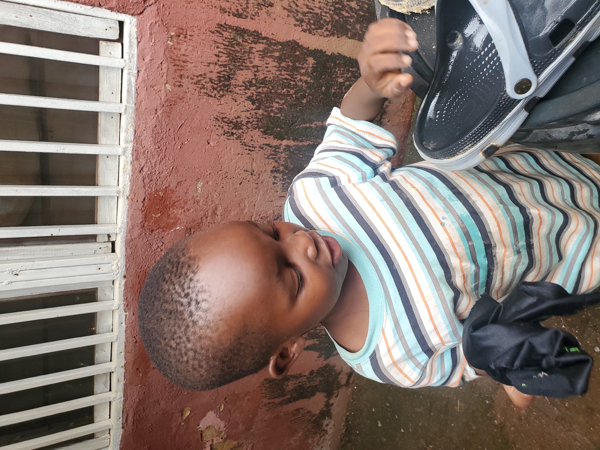 A young boy, appearing to be approximately 3-5 years old, with dark skin and short hair, is shown in a medium close-up shot outdoors. He is wearing a horizontally striped t-shirt with alternating bands of white, light blue, dark blue, orange, and grey. His head is tilted down as he looks at something out of frame to his right. In his left hand, he holds a crumpled piece of black fabric. His right hand is extended towards a black clog-style shoe with a light grey strap, which is resting on a dark object, possibly a bucket or a large shoe.
The background consists of a rough, reddish-brown textured wall, appearing to be stucco or concrete, with patches of dark, moss-like growth, suggesting a damp environment. Above the wall, a section of a white-painted window grill with vertical bars is visible. The ground beneath the boy is wet and appears to be dirt or muddy. The overall scene suggests a casual outdoor setting, possibly a backyard or alleyway, within the city of Jos, Nigeria, characterized by the building materials and the presence of water on the ground.