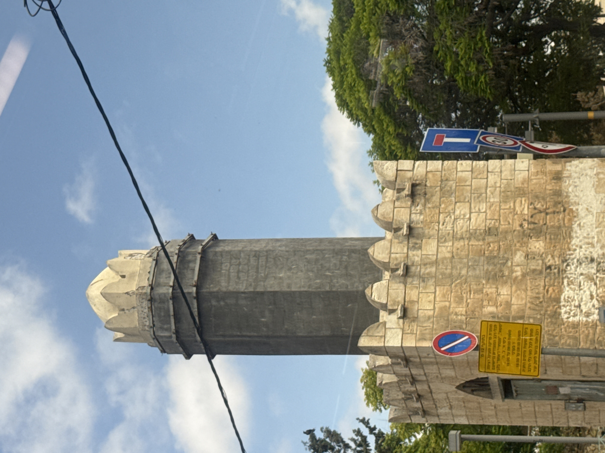 Here is a detailed description of the image:
The image shows a tall, stone tower rising into a blue sky with some wispy clouds. The tower has a distinctive design, featuring a cylindrical shaft that tapers slightly towards the top, and a decorative crown-like structure at its peak. The tower appears to be part of a larger complex, as a section of a stone wall is visible adjacent to it. The wall is constructed from light-colored stones.
The surrounding environment includes green trees and foliage, suggesting the tower is located in an area with vegetation. A street sign is also visible. A power cable runs diagonally across the left side of the frame. The OCR text appears to suggest that the parking in the area is permitted for all types of vehicles.
The architectural style of the tower and wall, along with the sign written in Hebrew, indicates that the photo was likely taken in Israel. Given the well-known historical and cultural significance of this building, it is most likely located in Jerusalem, Israel and NOT Tel-Aviv.