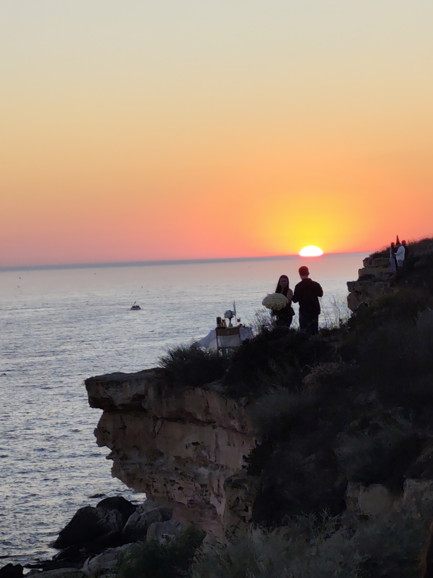 The image depicts a serene sunset scene on a cliff overlooking the sea. The sky is painted with warm hues of orange, pink, and yellow, transitioning to a soft gradient towards the top. The sun, a bright orb, is partially submerged below the horizon, casting a warm glow on the water. The sea is calm with gentle ripples, reflecting the colors of the sunset.
On the edge of the cliff, a couple stands silhouetted against the vibrant sky. The man is wearing dark clothing, and the woman, also in dark attire, is holding a large bouquet of white flowers. They appear to be engaged in a private moment, possibly a romantic proposal or a special celebration. Behind them, further up the cliff, a few other figures are visible, also silhouetted.
A small table with some items on it is set up near the couple, hinting at a planned event or setup. In the distance on the sea, a small boat can be seen, adding to the tranquil atmosphere. The cliff face is rugged and textured, with rocky outcrops and sparse vegetation. The overall mood of the image is peaceful and romantic, capturing the beauty of nature and a tender human interaction. The time of day is clearly sunset, and the weather appears to be clear and calm. The location cues of rugged cliffs and the sea suggest a coastal setting, possibly Aktau, Kazakhstan, known for its dramatic landscapes. There is no visible text in the image.