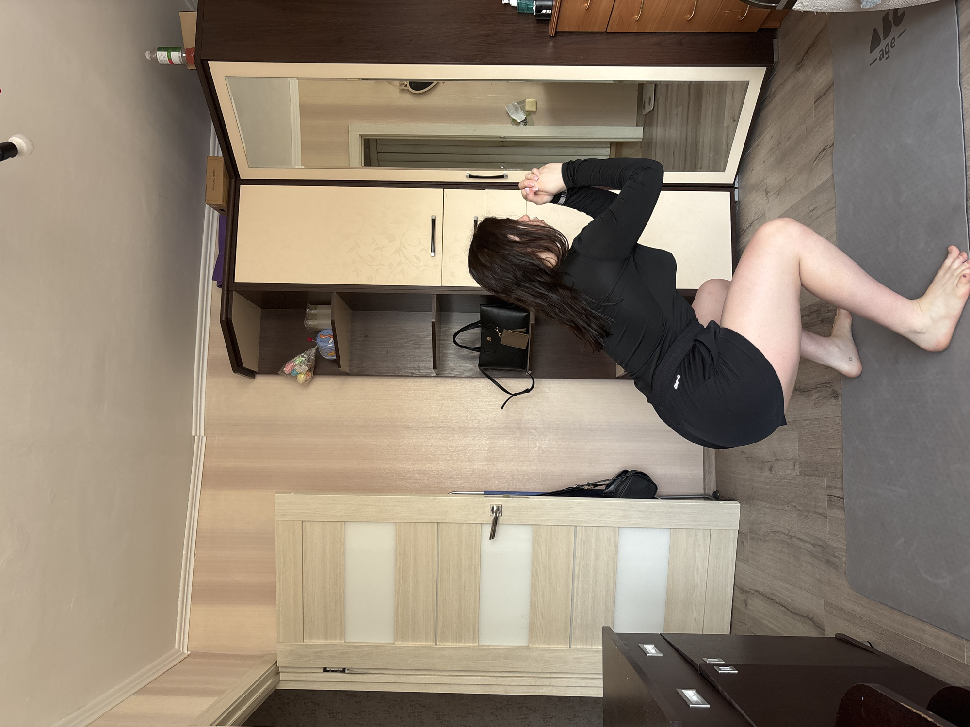 This image depicts an indoor scene within a residential room, located in Perm, Russia. The primary subject is a female individual, observed from her right side, engaged in a squat exercise on a grey yoga mat. She has long, dark hair and is barefoot, wearing a black long-sleeved athletic top and black shorts. A small heart-shaped tattoo is visible on the dorsal aspect of her left foot, near the ankle. Her hands are clasped together in front of her chest as she maintains the squat position.
The room features light brown wood laminate flooring and walls covered in vertically striped wallpaper of beige and pink hues. White crown molding runs along the ceiling, which is white and features a black track light fixture with a white bulb in the upper center.
On the left side of the frame, a dark brown dresser with silver drawer pulls is partially visible. Adjacent to it is a light-colored wooden door with five horizontal frosted glass panels and a silver handle, slightly ajar, from which a black handbag hangs.
Dominating the right side of the room is an L-shaped corner wardrobe unit. The left section of this unit has a dark brown frame and light beige paneled doors, with open shelves displaying small decorative items and a black handbag. The right section of the wardrobe consists of a full-length mirrored door, which reflects a partially open doorway, a light-colored wall with a heart-shaped decoration, and a white digital bathroom scale on the floor beyond. On top of the wardrobe, a purple object, a brown cardboard box (labeled "Cheers The Future"), and a plastic bottle are visible. To the far right, a section of a dark brown dresser with brass-colored handles and a clear pump bottle (likely hand sanitizer) are also present. The grey yoga mat on which the individual is exercising displays the text "ABF-age" in its lower right corner.