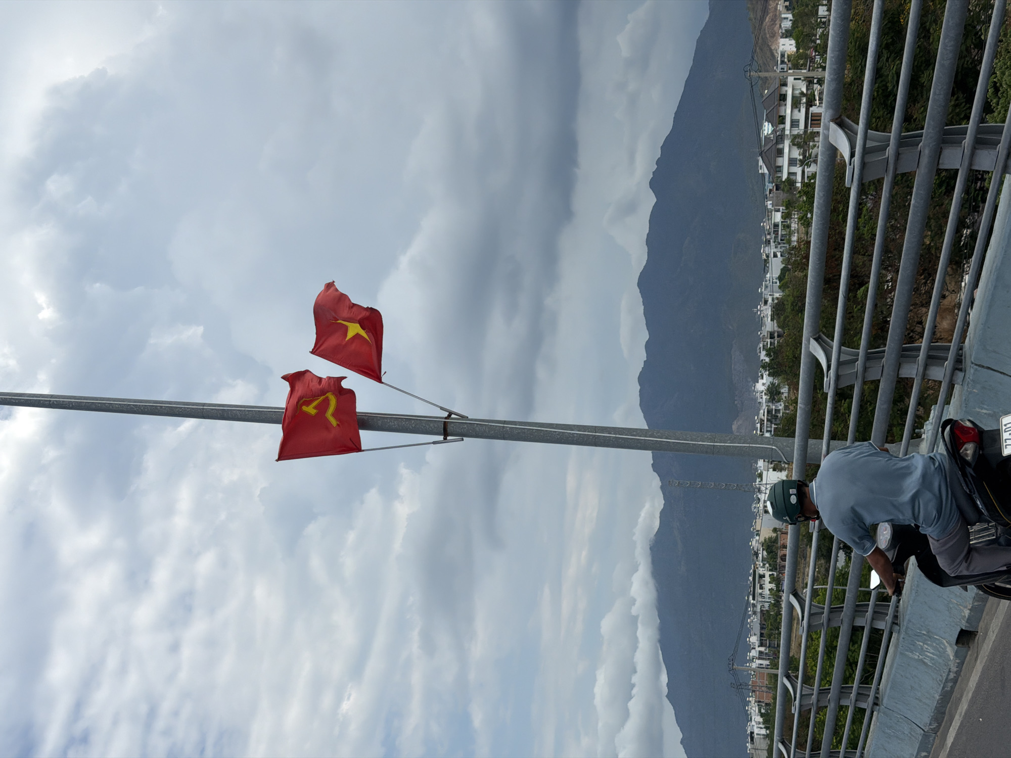 A medium shot reveals two Vietnamese flags mounted on a pole against a cloudy sky, adjacent to a mountainside partially obscured by haze. A man wearing a light blue shirt and green helmet is seated on a motorcycle on the right, appearing to be in motion on a bridge or elevated roadway. The setting is identified as Nam Nha Trang Ward, Vietnam, as indicated by text visible on the motorcycle's license plate. A railing runs alongside the road, and buildings are sparsely visible on the mountainside.