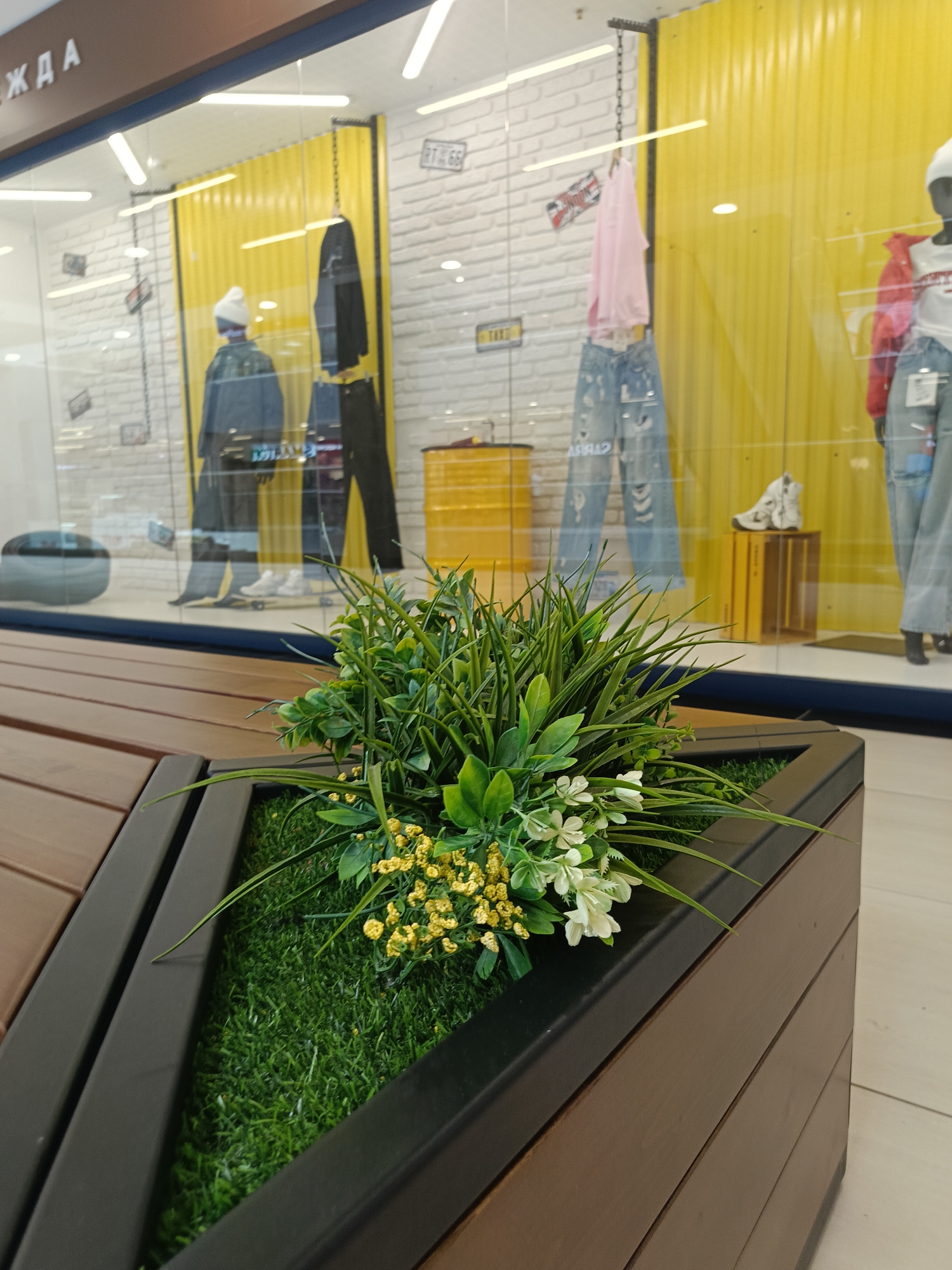 A close-up shot focuses on a planter filled with artificial grass, green foliage, and small white and yellow flowers, set within a wooden and black metal structure. The planter is positioned in front of a store window display, featuring mannequins dressed in casual clothing against a backdrop of yellow and white textured walls. A sign with Russian lettering, "ЖДА," is visible at the top of the window. Other items such as a yellow barrel, shoes, and various clothing items are also displayed. The flooring is light-colored, suggesting a retail or public space. The location is identified as the city of Izhevsk, Russia.