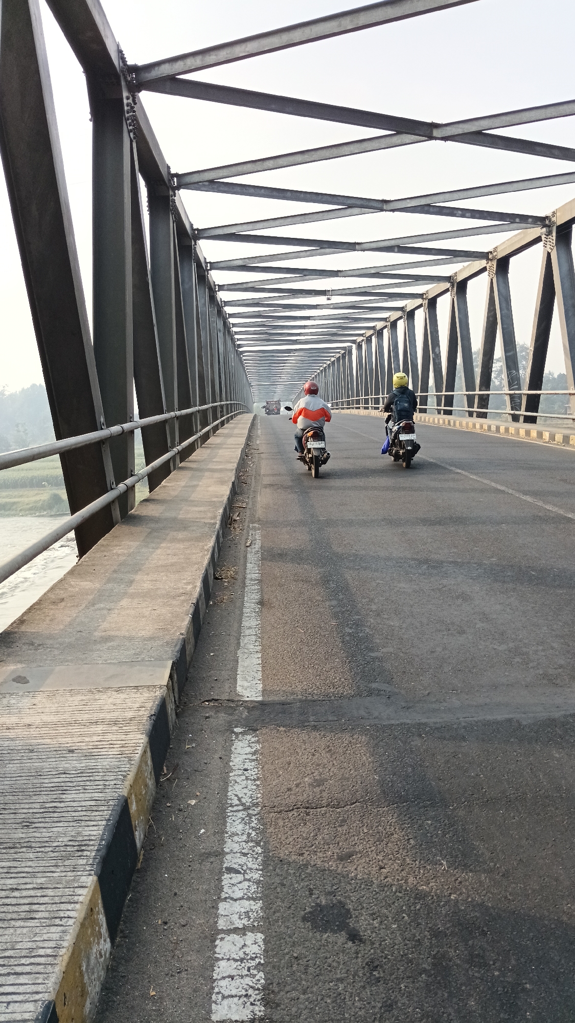 The image shows a view from a road bridge over a river in Mojo, Indonesia. Two people on motorcycles are traveling away from the camera on the paved road. The bridge has a dark grey steel truss structure. The sky is overcast. The river flows to the left of the bridge.