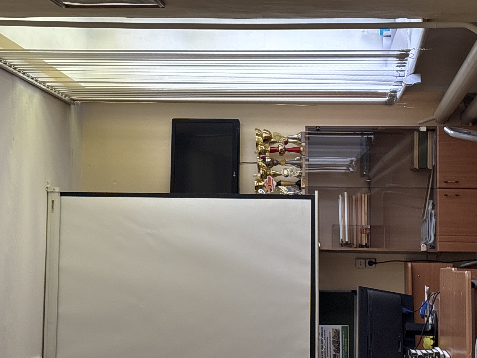 The image depicts a section of an interior space, likely an office or classroom. Dominating the foreground is a large whiteboard with a black frame. Behind the whiteboard, a flat-screen TV is mounted on the wall, next to shelves that hold a collection of trophies. Various other objects are present, including what appears to be a computer monitor, along with wooden cabinets and shelves. The ceiling features fluorescent lighting fixtures. The city and location are unknown.