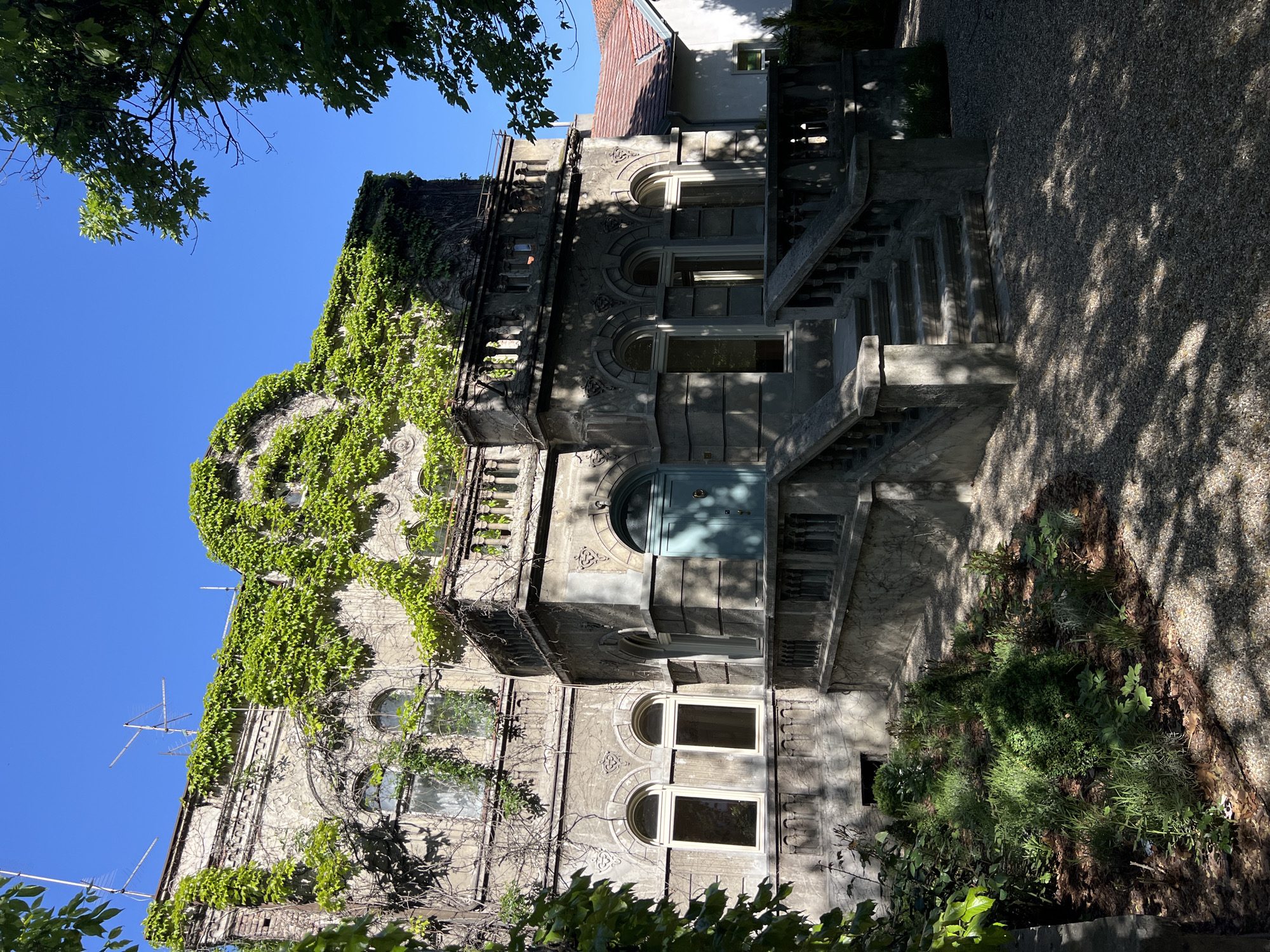 The image depicts a multi-story building in Belgrade, Serbia, constructed in a somewhat dilapidated, turn-of-the-century architectural style. The facade is predominantly grey and covered in climbing ivy, especially near the top of the building. The building features several arched windows and a light blue arched doorway on an upper level, accessible by a concrete staircase. There appear to be external stairs to different levels. The yard is covered in gravel and plant life. Shadows from trees and foliage are cast onto the ground. The sky is clear and blue. There is a television antenna affixed to the building on the left side.