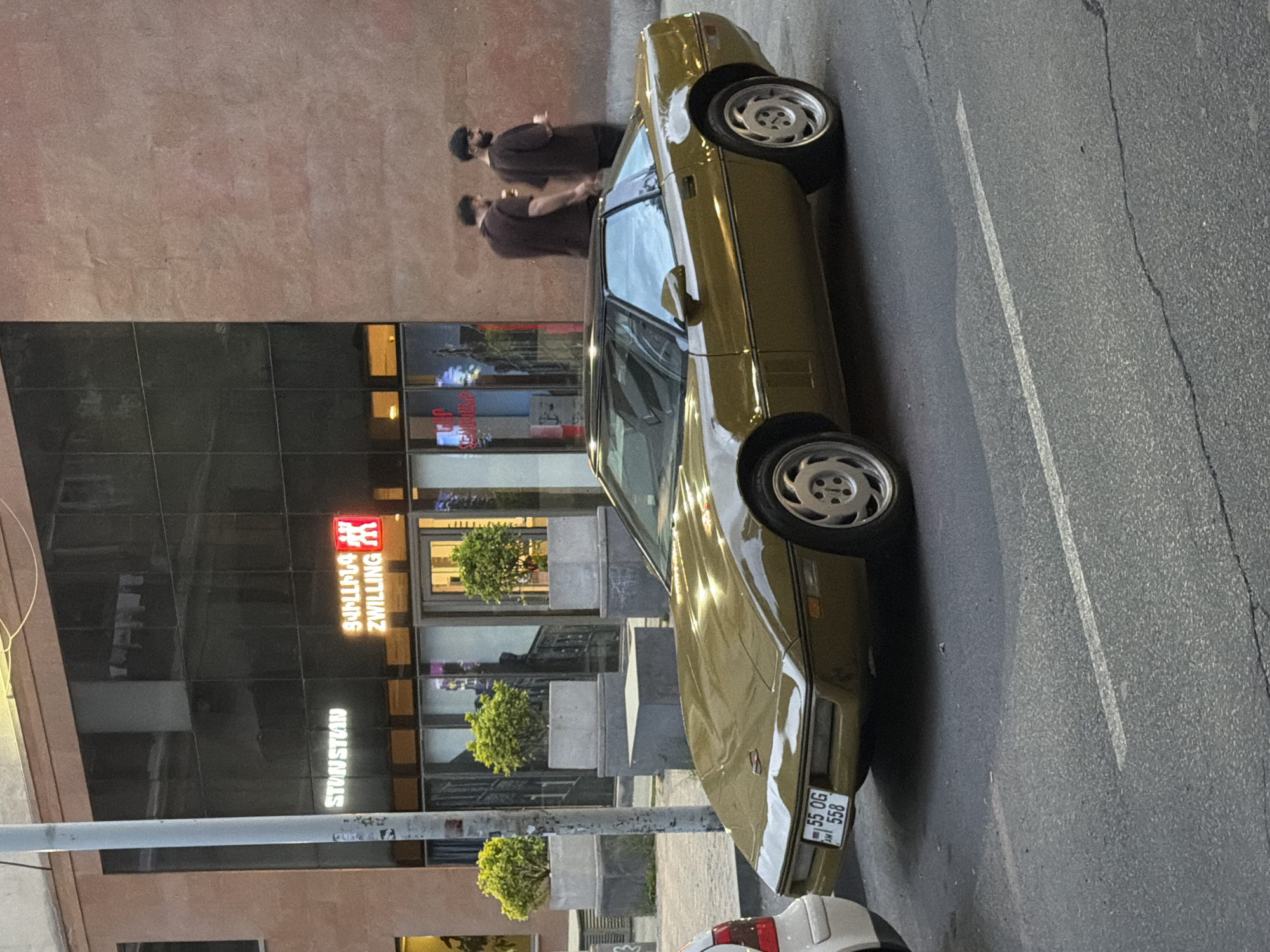 A gold-colored car is parked on a street in Yerevan, Armenia. The car is facing away from the viewer and slightly to the right. Two individuals are standing near the vehicle on the left side of the frame, near a building featuring the "Zwilling" logo and text in what appears to be Armenian. The architecture is modern, with large windows and a metallic structure, possibly a light post. The ground is asphalt, marked with white road lines. A portion of another white car is visible at the bottom right of the image, near the license plate of the gold car. The light suggests it is daytime.