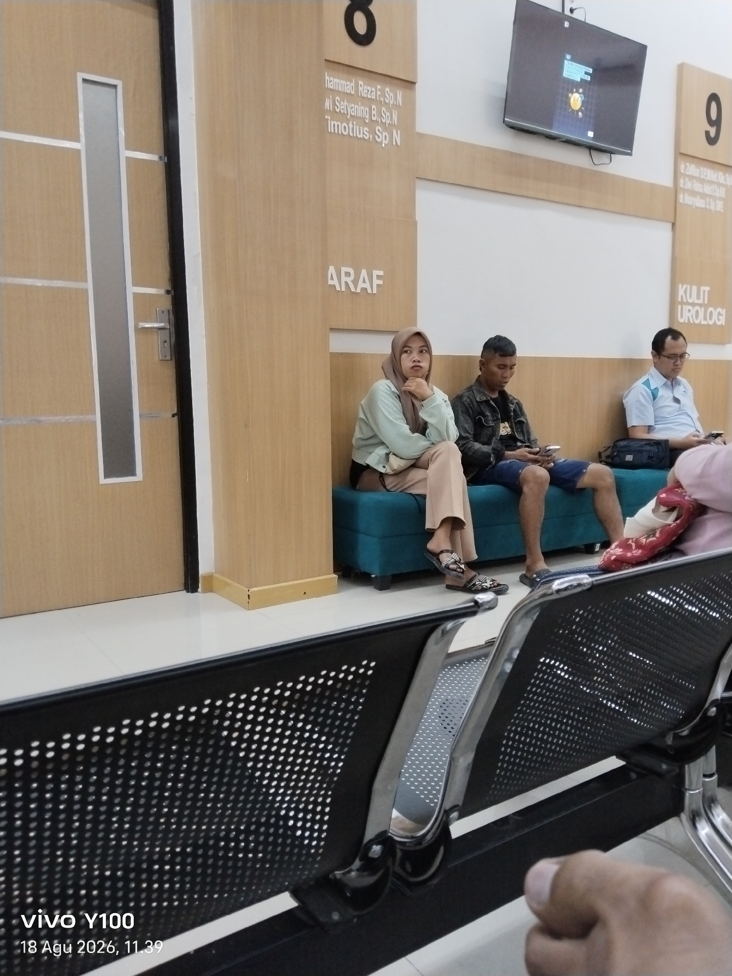 The image depicts a waiting area within a clinic or hospital, likely in Patokan, Indonesia, as suggested by the timestamp. In the foreground, a portion of a hand is visible, out of focus, suggesting the photo was taken from a seated position. The middle ground features a row of people sitting on a teal bench. From left to right, a woman wearing a light green hijab and pants is looking to her left. Next to her is a man in a dark denim jacket and blue shorts, looking down at his phone. To his right, a man in a light blue collared shirt and glasses is also looking at his phone. The background shows a hallway with light wood paneling. On the wall, there are signs with numbers and text, including "8" and "ARAF" on one side, and "9" and "KULIT UROLOGI" on the other. A television screen is mounted on the wall above the signs, displaying a graphic. The floor is tiled with white and grey squares. Chairs with perforated black seats and silver frames are present in the foreground and mid-ground.