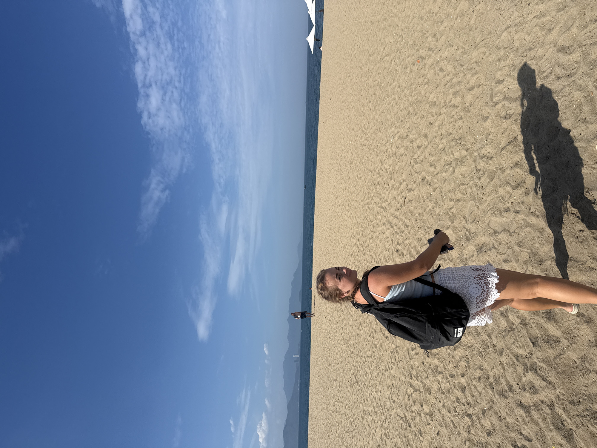 A young woman with blonde hair in a braid is standing on a sandy beach in Nha Trang, Vietnam, looking over her shoulder at the camera. She is wearing a light blue tank top and white crochet shorts, with a black backpack on. She is holding a dark-colored object in her right hand. The beach is wide and sandy, with gentle waves and the blue ocean stretching to the horizon. In the distance, there are mountains and a clear blue sky with scattered clouds. Two white umbrellas are visible on the beach further down.