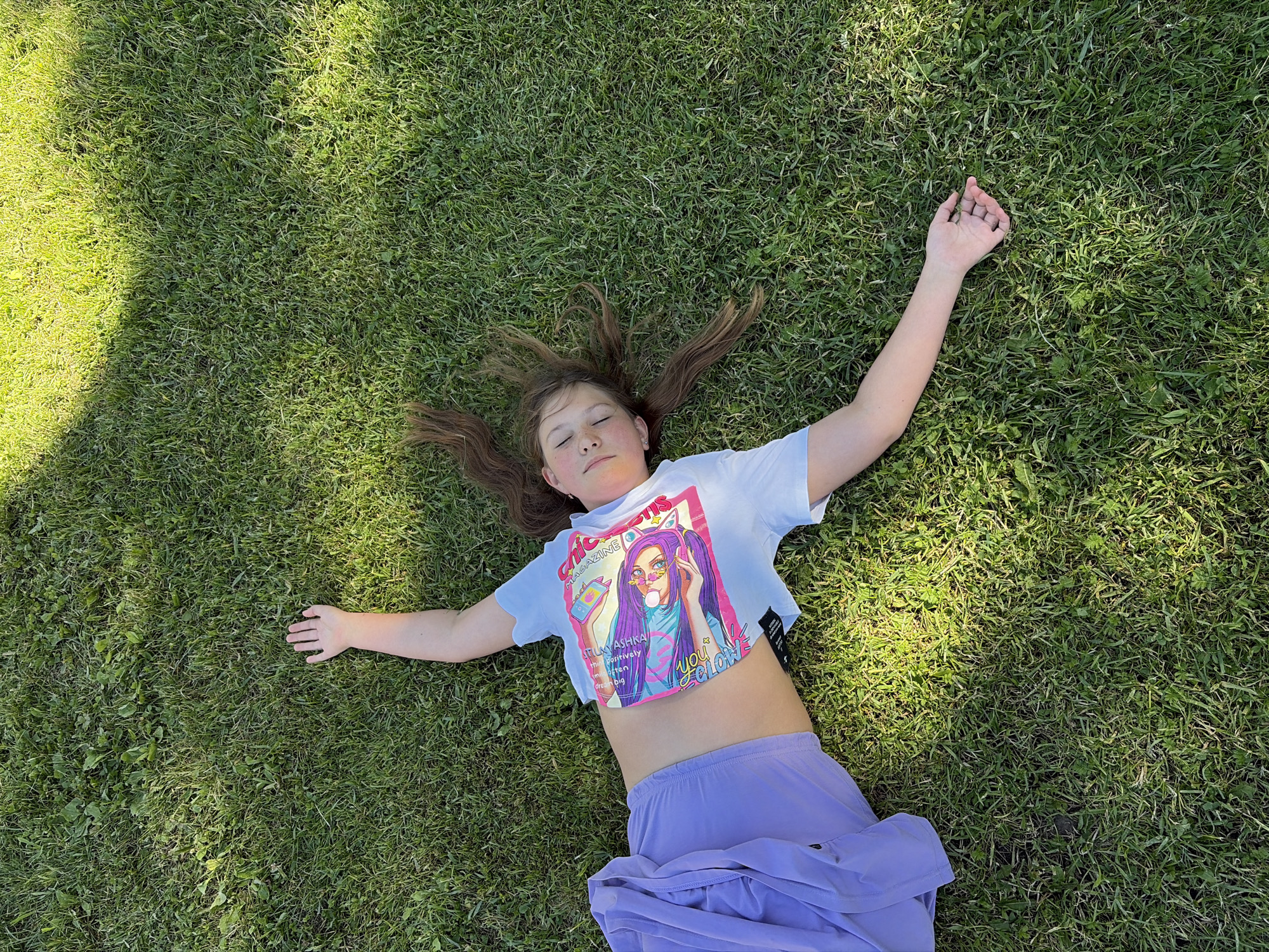 The image shows a young girl lying on her back on a grassy lawn. She has her arms and legs spread out, with her eyes closed and a relaxed expression on her face. Sunlight filters through the grass, casting shadows on the ground and highlighting the texture of the lawn. The girl is wearing a white cropped t-shirt with a colorful graphic print on the front, and light purple, flowing pants. Her brown hair is spread out around her head.
The graphic on the t-shirt features an illustration of a person with purple hair wearing headphones and sunglasses, and appears to be styled like a magazine cover. Text visible on the graphic includes "CHICONS", "TELMA ASHKA", and "you color".
The scene suggests a warm, sunny day, possibly in the afternoon, given the angle of the sunlight and the shadows. The presence of a manicured lawn indicates an outdoor setting, such as a park or a backyard. The overall mood is one of peace and tranquility, as the girl appears to be enjoying a moment of rest or contemplation in nature. The location context provided is Saint Petersburg, Russia, which can be inferred as the geographical setting for this image.