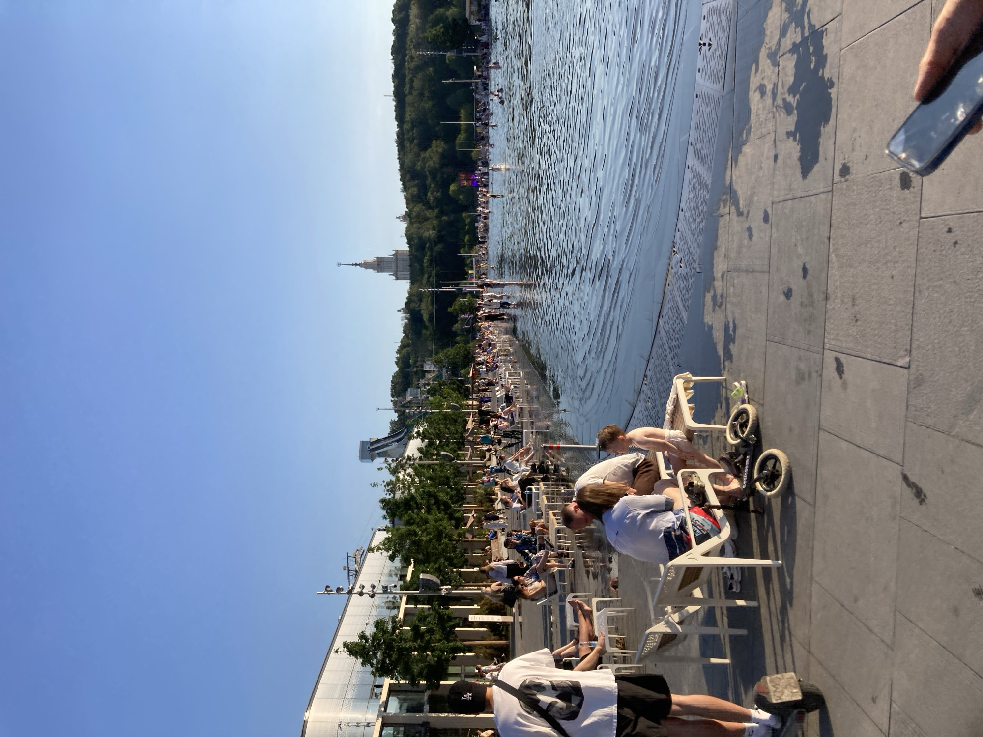The image depicts a wide shot of a large public fountain area in Moscow, Russia, under a clear blue sky. In the foreground, several people are seated on white chairs along the edge of the fountain, some with their feet in the shallow water. A man on the left is standing with his back to the camera, wearing a white t-shirt and black shorts, with a backpack slung over his shoulder. A child in a diaper is sitting on a bench, facing away from the camera. Further into the scene, the fountain extends into a larger body of water where numerous people are wading and playing. In the background, a modern building with a curved glass facade and trees line the edge of the fountain area. A tall, distinctive building with a spire, likely the Moscow State University, can be seen in the far distance, set against a wooded hillside. The ground around the fountain is paved with large gray tiles, some of which are wet from the water. The overall atmosphere is relaxed and recreational, suggesting a warm day at a popular urban park.