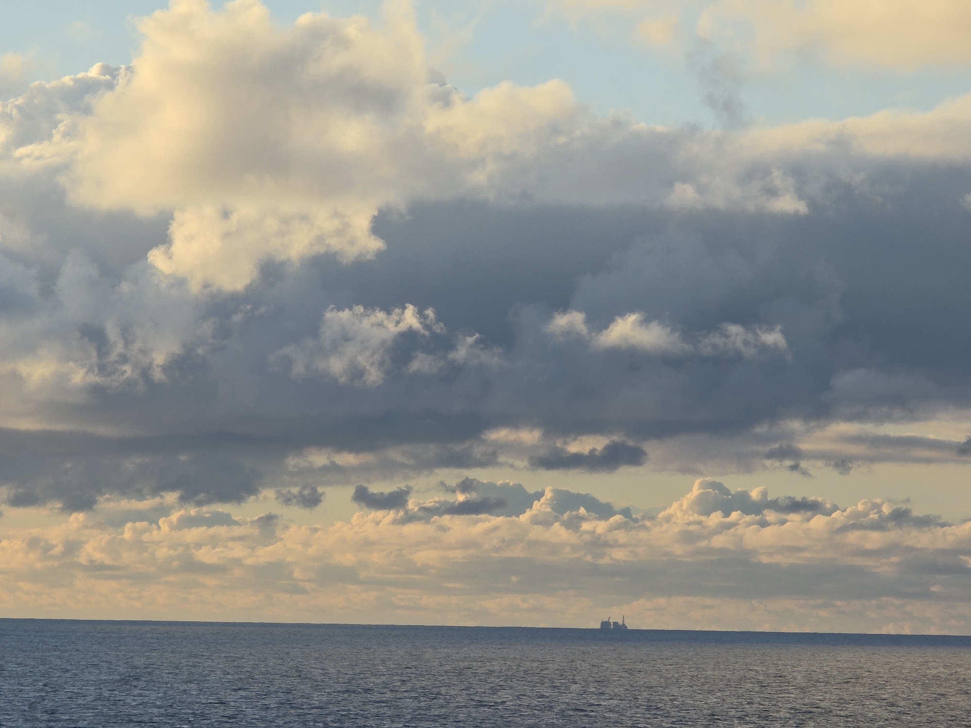 The image depicts a body of water, possibly an ocean, with a cloudy sky above. In the distance, there is a structure on the water, potentially an oil rig or a large ship. The clouds are layered, with some being illuminated by the setting or rising sun, creating a soft, golden glow. The environment is serene and vast, dominated by the elements of water and sky. The location is unknown.