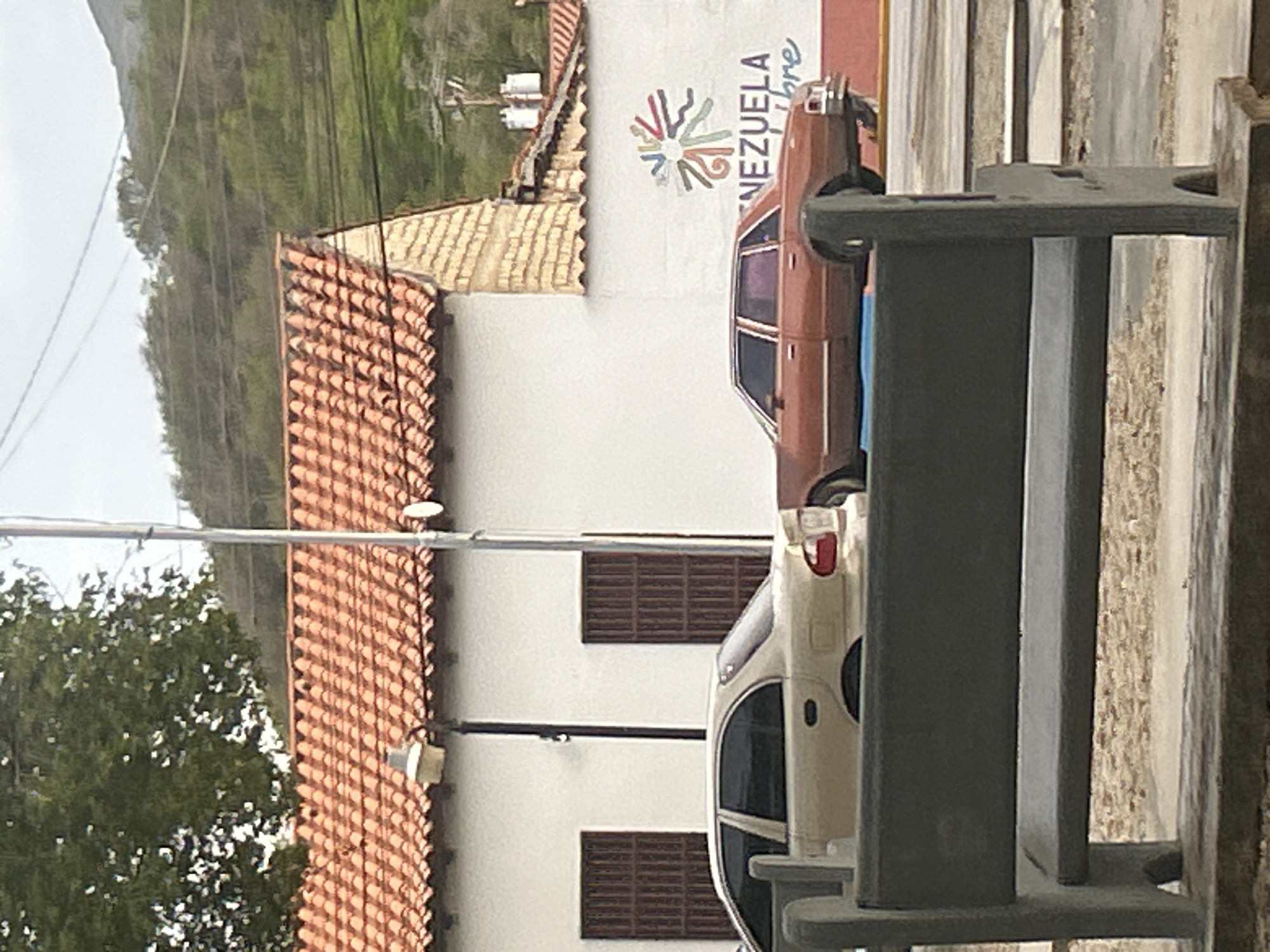 A light-colored sedan is parked next to a red sedan on a street in front of a building. The building has a red tile roof and white walls. The red sedan has a mural painted on the wall behind it that says "VENEZUELA LIBRE". A bench is in the foreground, with a blurred view of the street behind it. The background shows a hillside covered in trees. The scene is likely in Santa Ana, Venezuela.