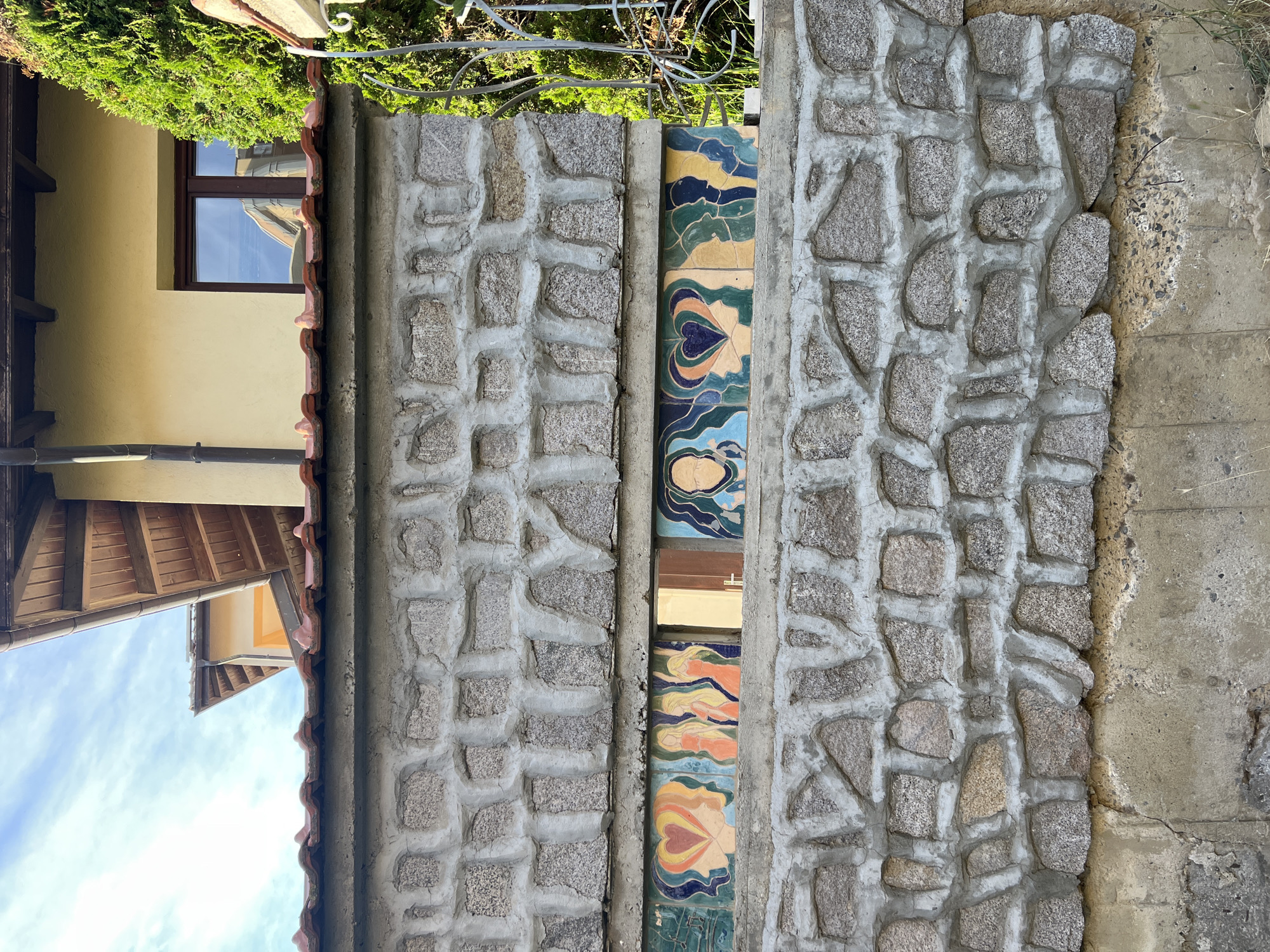 The image shows a section of a wall constructed with rough, grey stones and concrete. In the center of the wall, there is a vertical strip of colorful decorative tiles featuring abstract patterns and stylized human figures. The tiles display a palette of blues, greens, yellows, oranges, and pinks. To the left of the stone wall, there is a portion of a building with a yellow facade and a wooden-shingled roof. A window with a dark frame is visible on the yellow wall, reflecting a blue sky with some clouds. To the right of the stone wall, a rough concrete surface is visible. The scene appears to be outdoors, with green foliage at the top of the image, suggesting a daytime setting. The overall impression is of a rustic and artistic architectural detail.