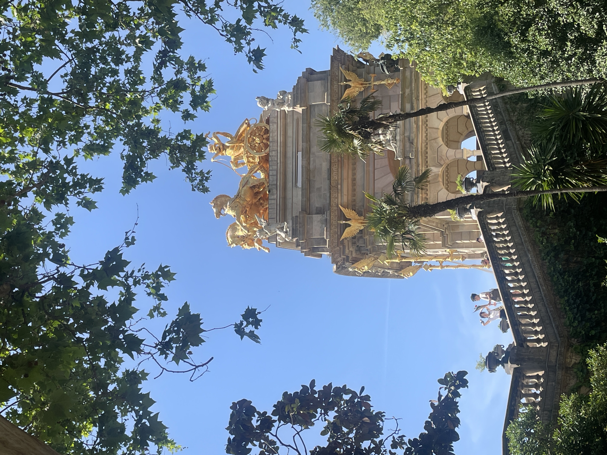 The image captures a low-angle view of the Cascada Monumental in Parc de la Ciutadella, Barcelona, Spain. The architectural structure is partially visible through the surrounding trees, displaying golden sculptures of horses and chariot riders atop the monument. Palm trees are positioned near the structure. Part of the staircase, with people standing on it, can also be observed on the side of the image. The sky is blue and clear, indicating a sunny day. Branches and leaves of trees frame the monument.