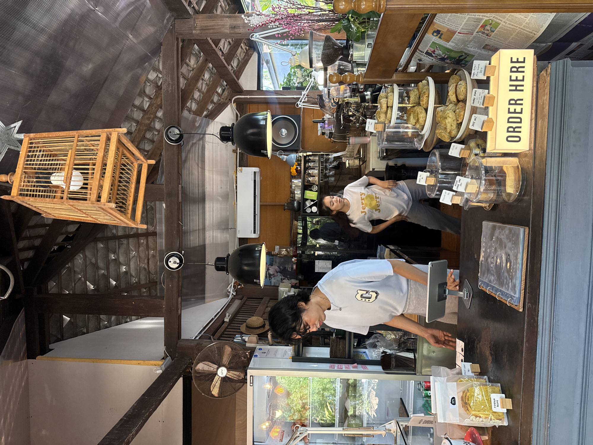 The image shows the interior of a cafe in Ko Samui District, Thailand. Two baristas, a man and a woman, are working behind a counter. The man, wearing a light blue t-shirt with a graphic on the front and grey pants, is standing on the left and appears to be interacting with a tablet on a stand. The woman, wearing a white t-shirt with a graphic and grey pants, is standing on the right and appears to be looking down and to her left. The counter displays baked goods in glass cases, a menu book, and various signs. Behind the counter, shelves are stocked with bottles and other items, and a coffee machine is visible. The cafe has a rustic aesthetic with exposed wooden beams, a pitched roof, and decorative lighting fixtures, including a wooden birdcage hanging from the ceiling. A large fan is mounted on the wall to the left. Natural light streams in from an unseen source.
