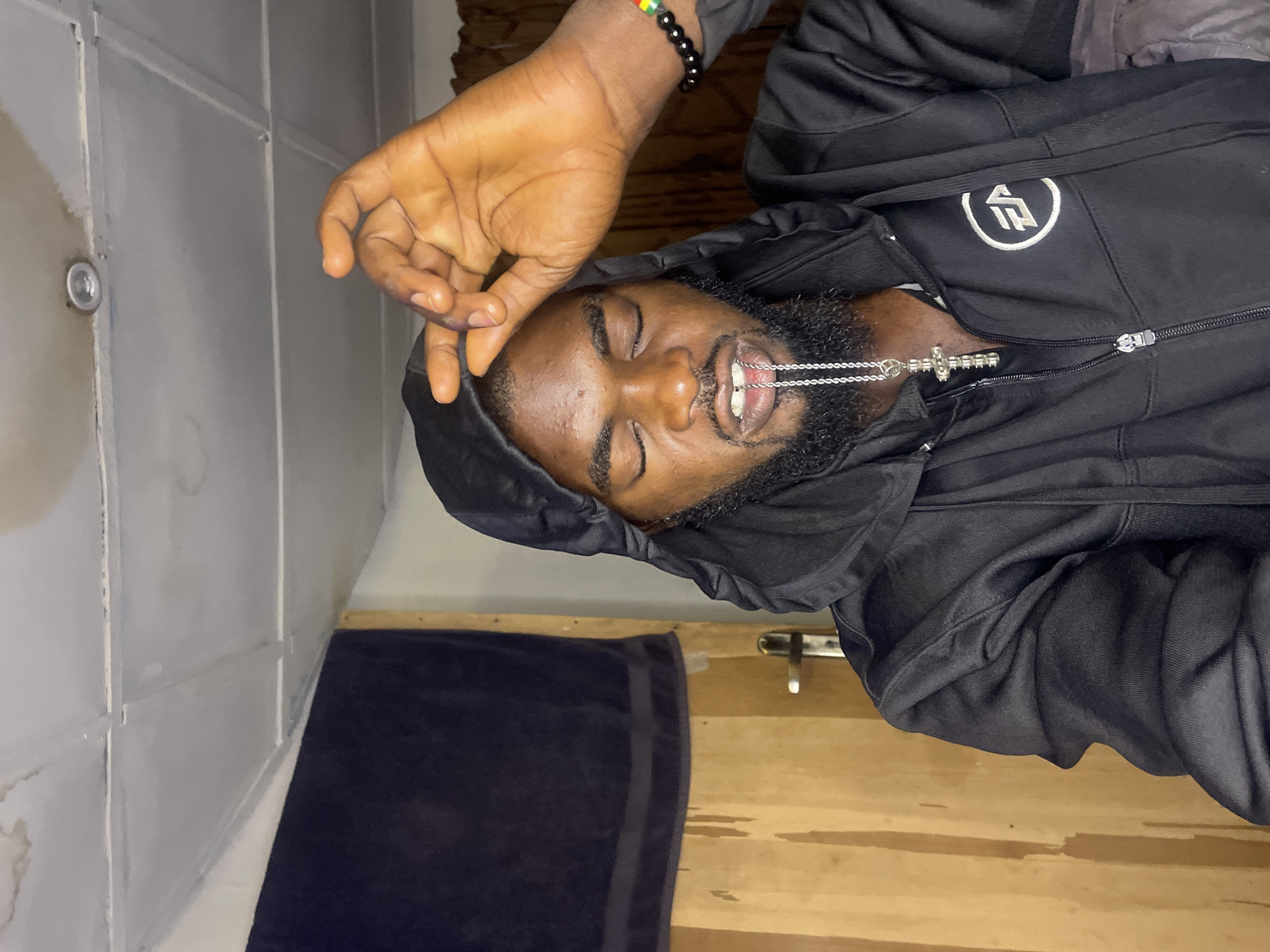A man with dark skin, a beard, and closed eyes is lying down, wearing a black hooded jacket with a silver cross necklace in his mouth. He is also wearing a black bracelet with green, yellow, and red beads on his left wrist. The background appears to be an indoor space with a white tiled wall on the left, a black towel hanging on a wooden surface behind him, and a glimpse of a wooden floor. The photograph is taken at an odd angle and is potentially sideways. The location is not specified, so it is impossible to locate it in the city of Jos, Nigeria.