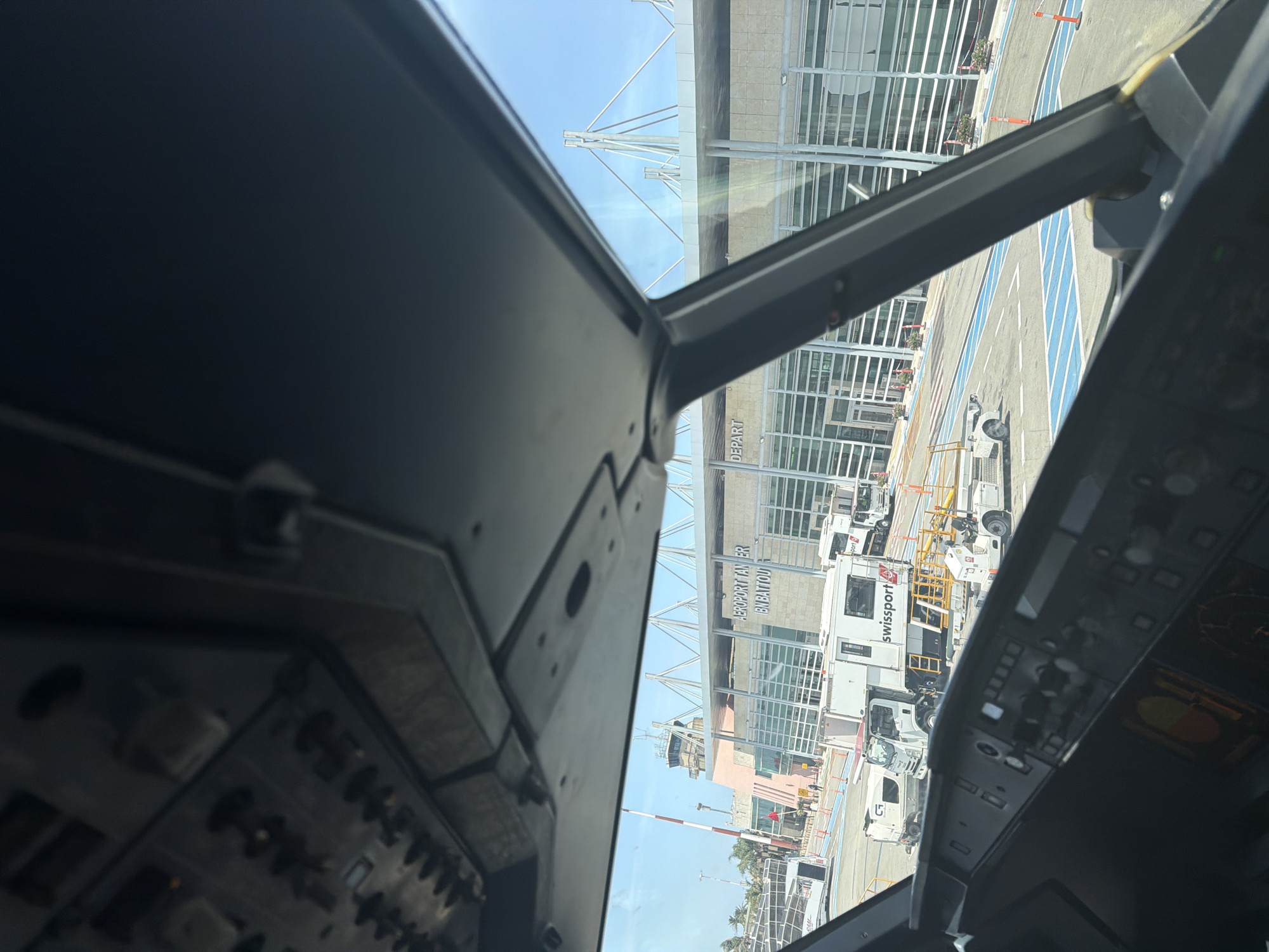 The image presents a view from the cockpit of an aircraft parked at Tangier Ibn Battouta Airport in Tangier, Morocco, on a clear, sunny day. The upper section of the aircraft's overhead panel and the top of the main instrument panel, with some illuminated displays, frame the scene. Directly ahead, the modern terminal building is visible, featuring large glass facades and a distinctive roof structure with white support beams. The name "AEROPORT TANGIER IBN BATTOU" is clearly displayed on the building's exterior, along with "DEPART" to the right. To the left of the main terminal, a control tower sits atop a salmon-pink structure, accompanied by a red and white striped antenna. On the tarmac, various ground support vehicles are positioned. A prominent white Swissport truck, with its red logo, is situated in the mid-ground, alongside other ground handling equipment and what appears to be a baggage cart. Blue-marked taxi lines and orange traffic cones delineate sections of the tarmac. A large passenger bus is partially visible on the far left. Distant palm trees suggest a warm climate. A few small, indistinct figures of people are discernible near the terminal building.