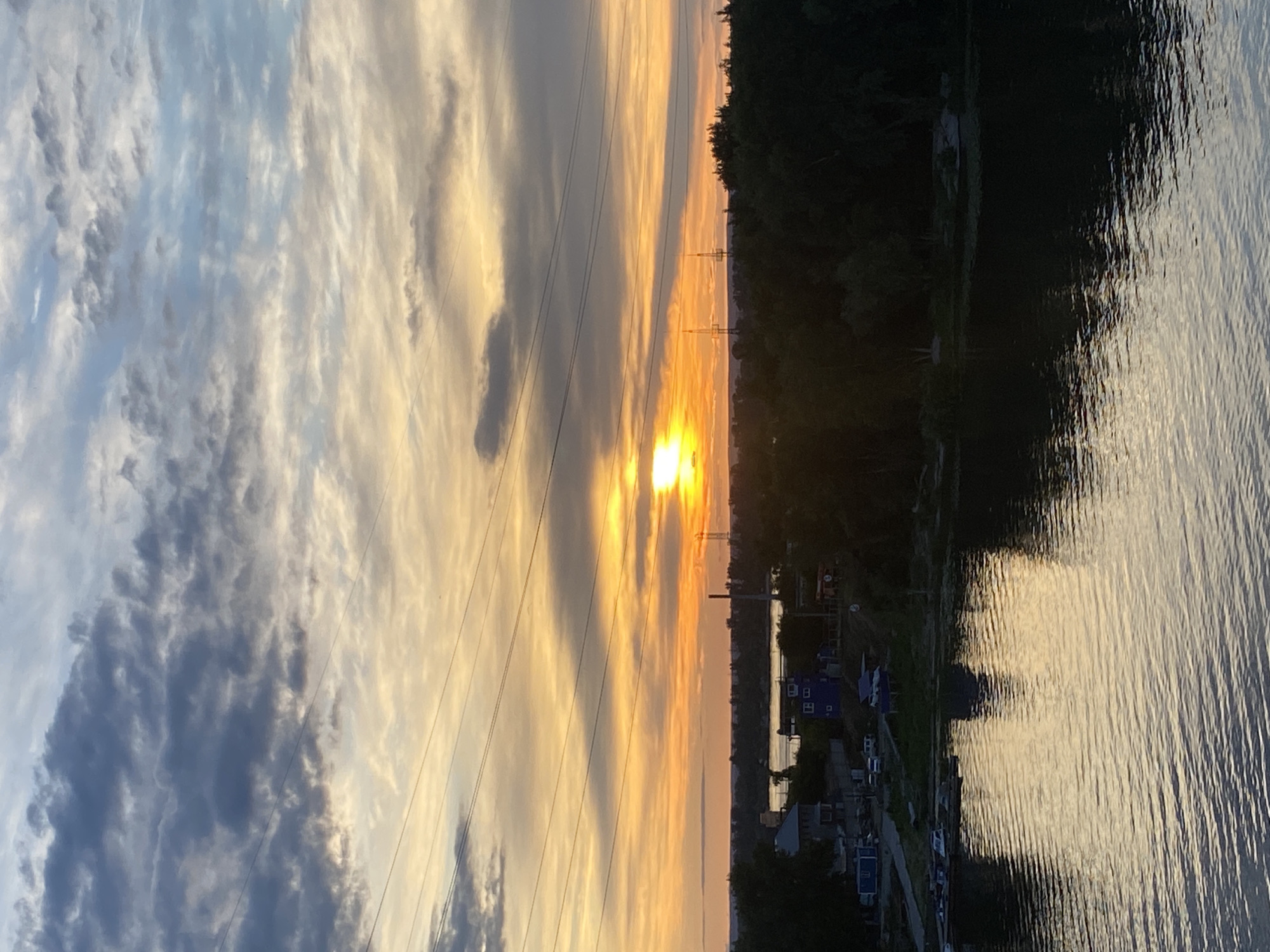 The image depicts a sunset over a body of water in Novokuybyshevsk, Russia. The sky is filled with dramatic clouds, tinged with orange and yellow hues from the setting sun, which is partially obscured by them. Silhouetted against the sky are power lines stretching across the frame.
In the middle ground, a treeline separates the water from a strip of land with some buildings and what appear to be industrial structures, possibly including cooling towers or transmission towers, indicating an urban or industrial area. The water in the foreground is calm, reflecting the colors of the sky. Some small boats are visible docked on the far side of the water, near a small cluster of houses or buildings. The overall scene is peaceful, with the setting sun casting a warm glow over the landscape.