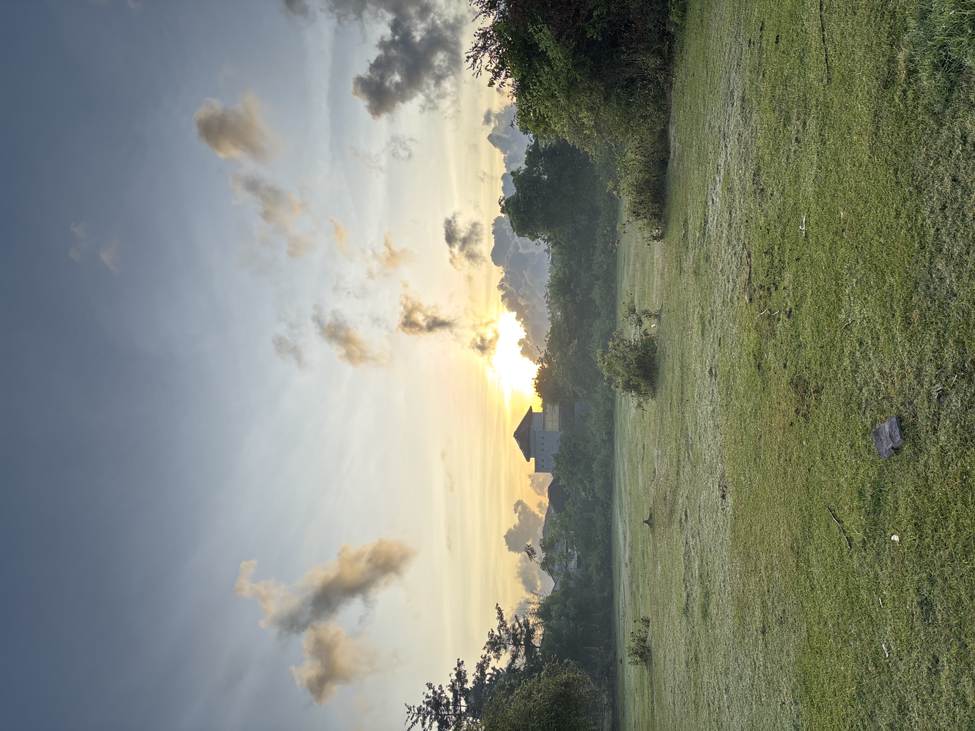 The image depicts a landscape at sunrise in Kuta Selatan, Indonesia. A building silhouette is visible in the background, centered in the frame and partially obscured by trees. The sun is positioned directly behind the building, casting a golden glow. A field of green grass occupies the foreground, with darker vegetation along the right edge. The sky exhibits a mix of light blue and gray clouds.