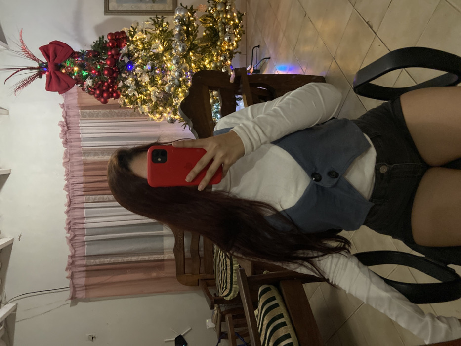 A person sits on a chair in front of a Christmas tree, taking a selfie with a red phone case. The individual wears a white long-sleeved shirt under a blue vest and black shorts. The Christmas tree is adorned with lights and ornaments, topped with a large red bow. Wooden furniture, striped cushions, and patterned curtains are visible. The lighting is soft, and the overall scene suggests a festive, indoor setting.