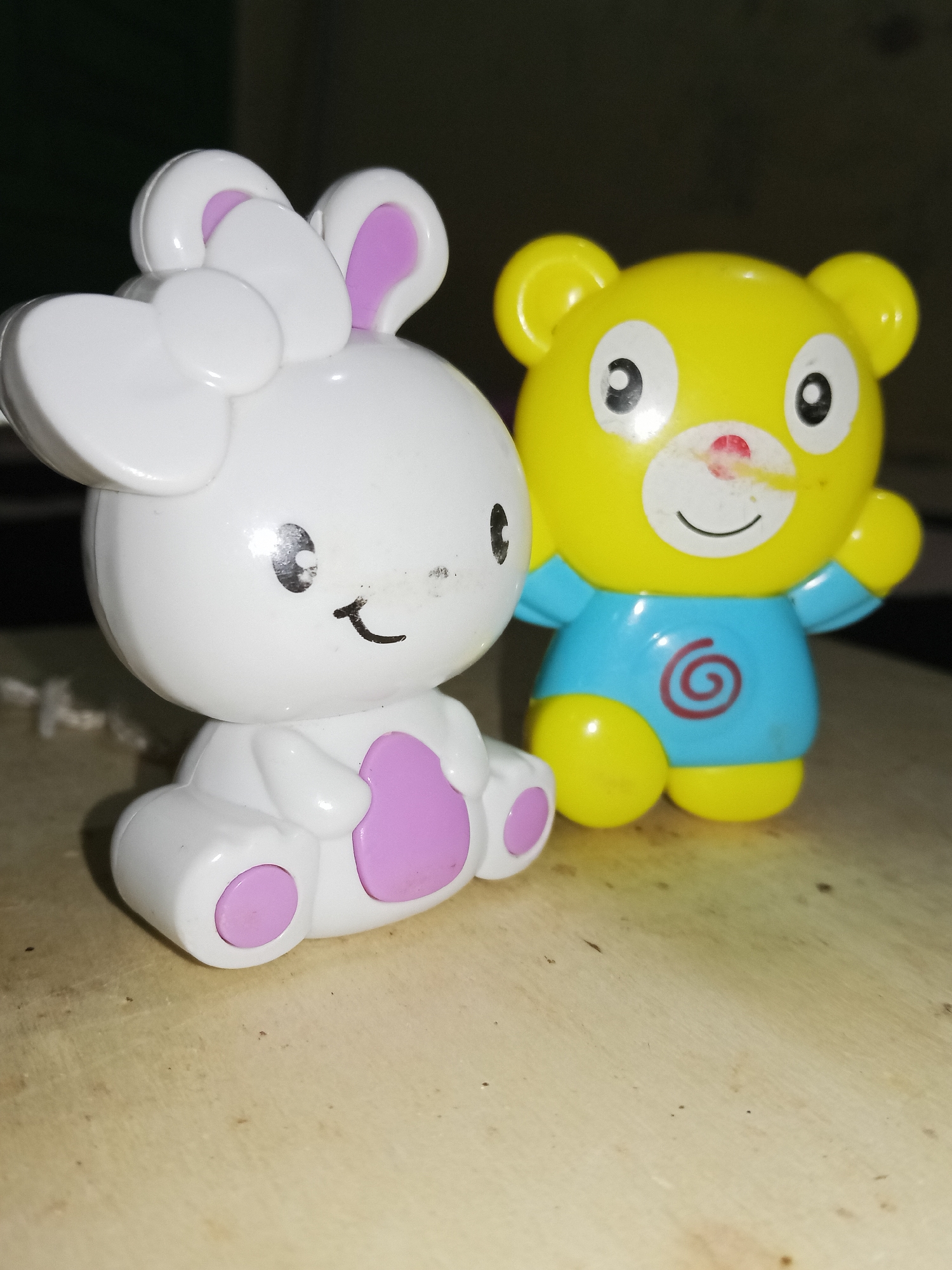 The image depicts two plastic toys on a wooden surface. On the left, a white bunny-like figure is seated, adorned with a bow and pink accents. To the right, a yellow bear-like figure stands, sporting a blue shirt with a red swirl. No people are visible. The specific location in Lhokseumawe, Indonesia is not discernible from the image.
