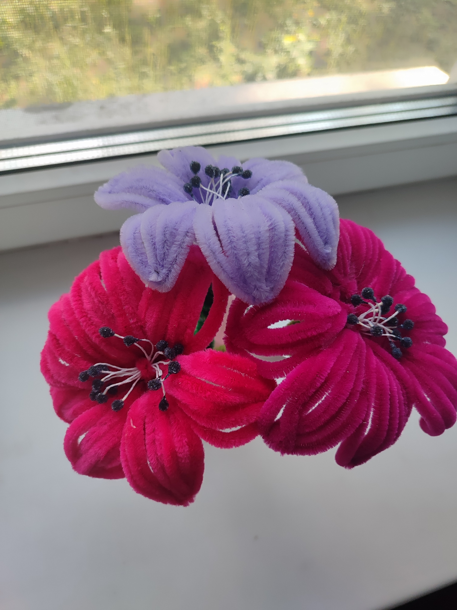 Three artificial flowers, two bright pink and one lavender, are arranged on a white windowsill. The flowers are made of fuzzy material and have black stamen. Behind the flowers, a blurred window shows a green, leafy outdoor scene. The windowsill is clean and well-lit. The overall impression is one of simple, decorative display.