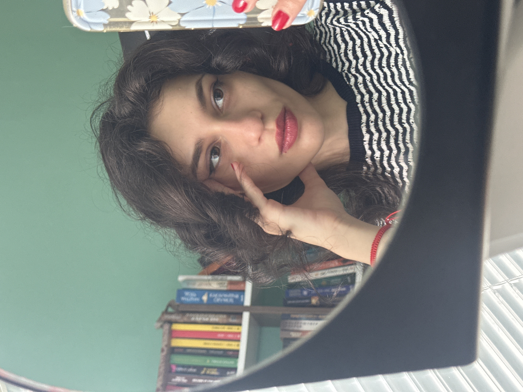 A young woman with dark, wavy hair and red lipstick is taking a mirror selfie. She is wearing a black and white striped sweater. Her left hand is resting on her cheek, and she has bright red nail polish. In the background, a bookshelf filled with books is visible against a teal-colored wall. The image is taken indoors, suggesting a home environment. There is no discernible location in Sofia, Bulgaria.