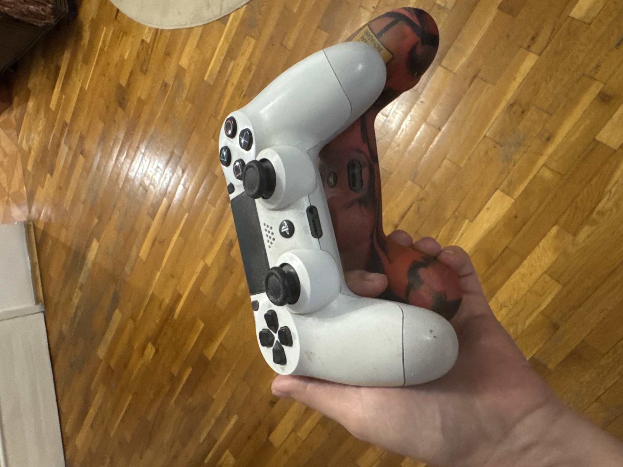 In the image, a person's hands are holding two video game controllers against a polished wooden floor. One controller is white and appears to be a PlayStation DualShock 4 controller, with its iconic button layout and analog sticks visible. The other controller is red and black, with a distinctive pattern that suggests it might be a custom-skinned controller or a different model. The controllers are positioned in a way that implies they are being held for gaming or demonstration.
The setting is indoors, with a focus on the wooden floor which has a warm, polished sheen, reflecting light. The lighting suggests an indoor environment, possibly during the day or with artificial lighting. The background is out of focus, but hints of furniture and a rug are subtly visible, suggesting a living space. There is no clear indication of the time of day or weather conditions. The emotions of the person holding the controllers are not discernible from the image.
One of the controllers has visible text on it. A label on the red and black controller contains text that appears to read "BIOHAZARD". The overall impression is that of a casual, domestic setting where video gaming is taking place. The Plovdiv, Bulgaria location context is not directly evident from visual cues within the image itself.