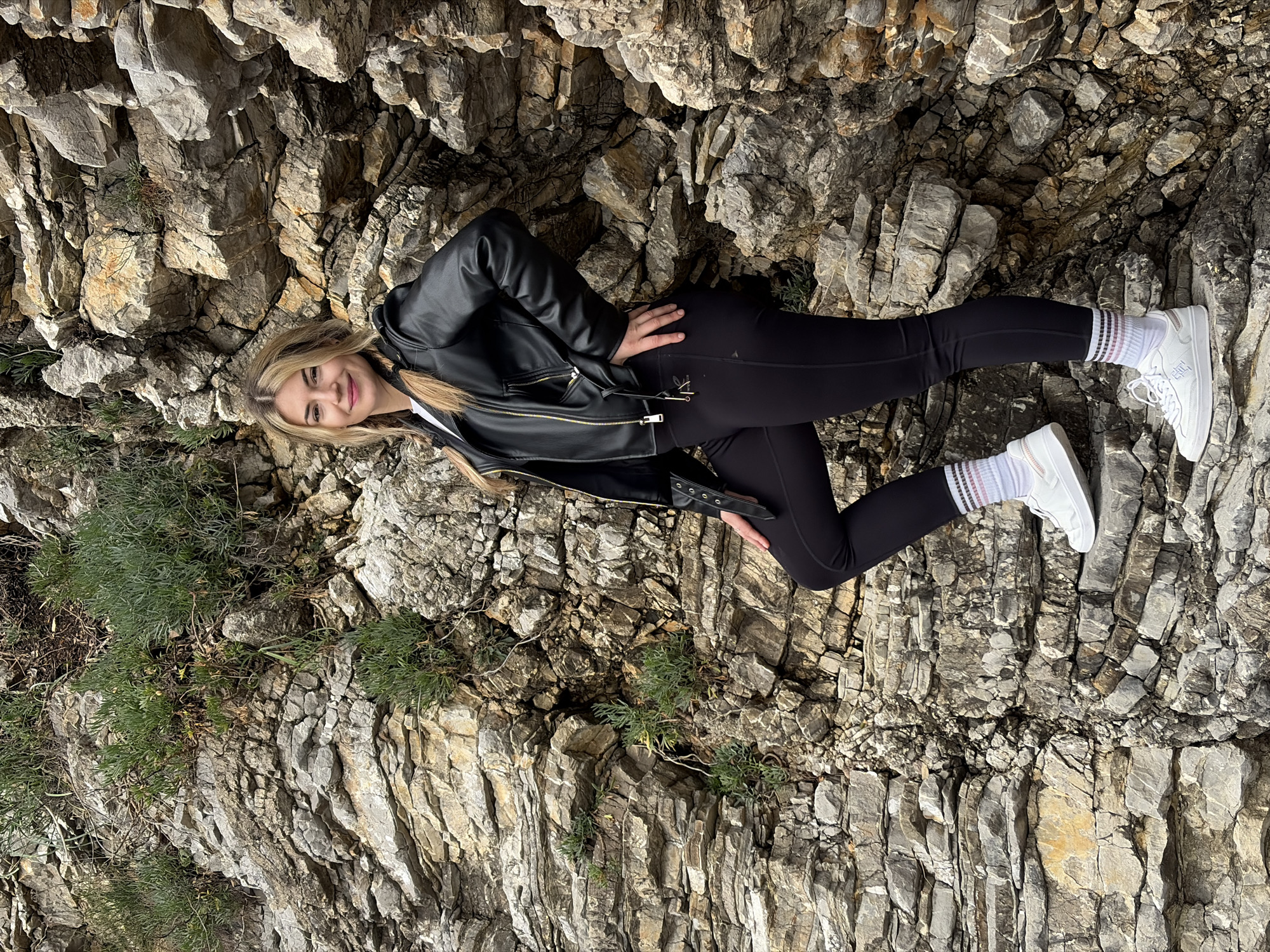 A young woman is posed on a rocky hillside in Budva, Montenegro. The geological formation is a light gray, layered rock face with crevices filled with small green plants. The woman is wearing a black leather jacket, a white top underneath, black leggings, white socks with black and pink stripes, and white sneakers. She is leaning back with her left hand on her hip and her right hand resting on the rock beside her. The scene is in daylight.