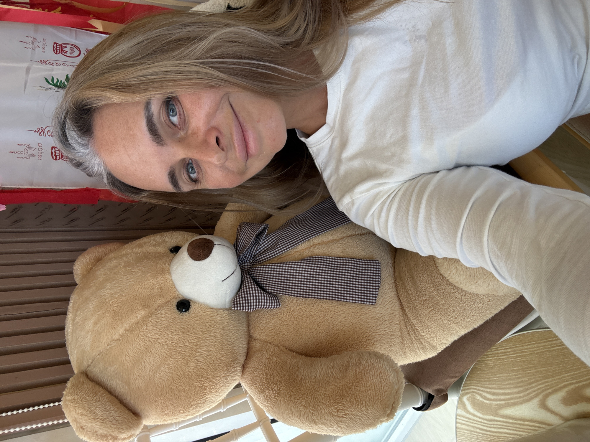 A woman with blonde hair and blue eyes smiles at the camera, seated next to a large tan teddy bear. She wears a white long-sleeved shirt. The teddy bear has a brown and white checkered ribbon around its neck. They are in an indoor setting with a wooden slatted wall and some decorations visible in the background. The image appears to have been taken in Ko Samui District, Thailand, based on the presence of religious banners commonly found in Thailand.