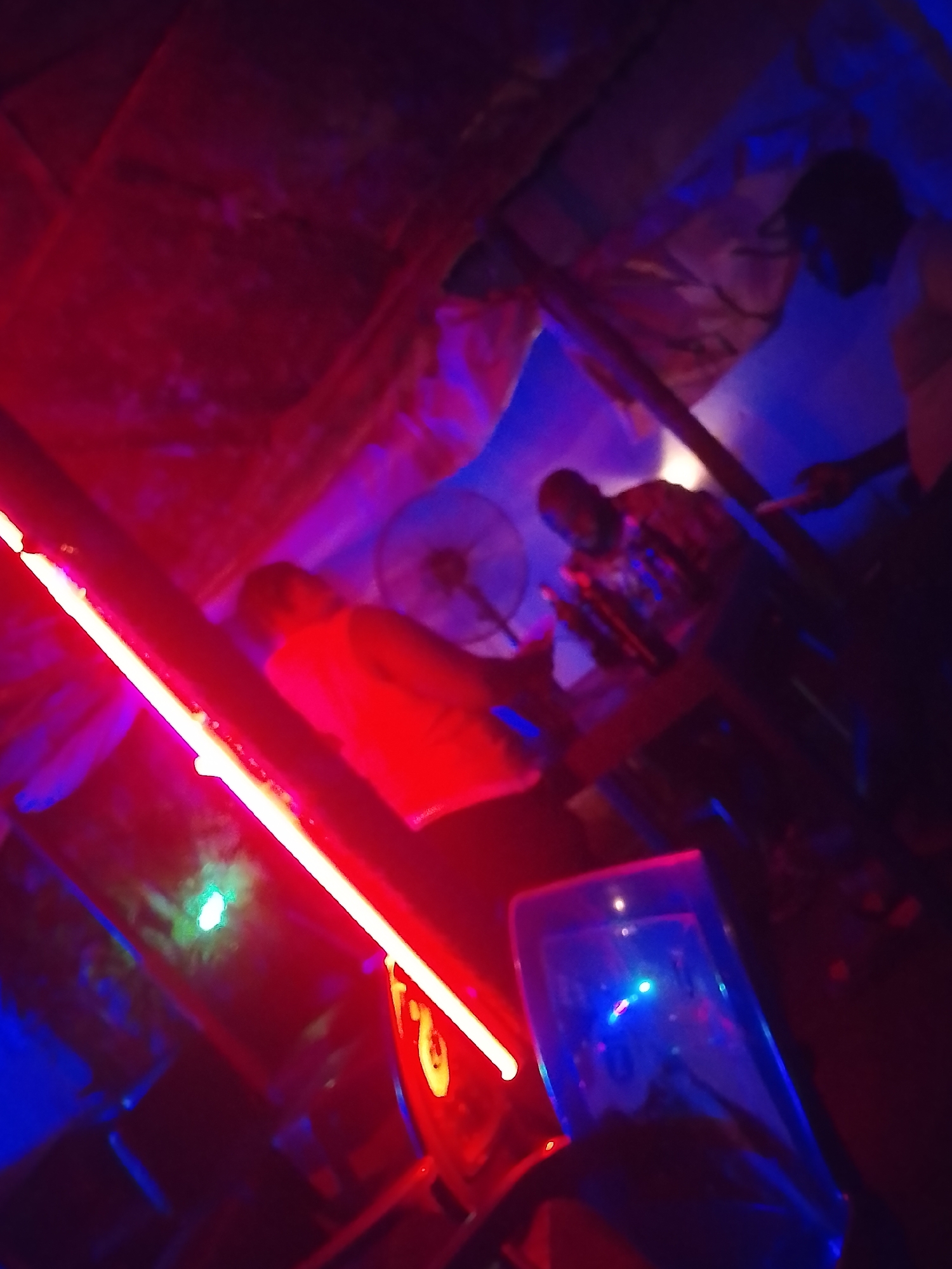 The image, taken in an unidentified location in the city of Unknown, Unknown, shows a poorly lit interior space under what appears to be a rudimentary ceiling. Red and blue lighting dominates the scene. Several people are visible, although details are obscured by the lighting. One person is wearing a red shirt, and another is wearing a patterned shirt. A fan is visible in the background. In the foreground, there is a plastic cup or container illuminated in blue light, positioned on a surface. A red light source is also in the foreground. The setting appears to be a bar or informal gathering space.