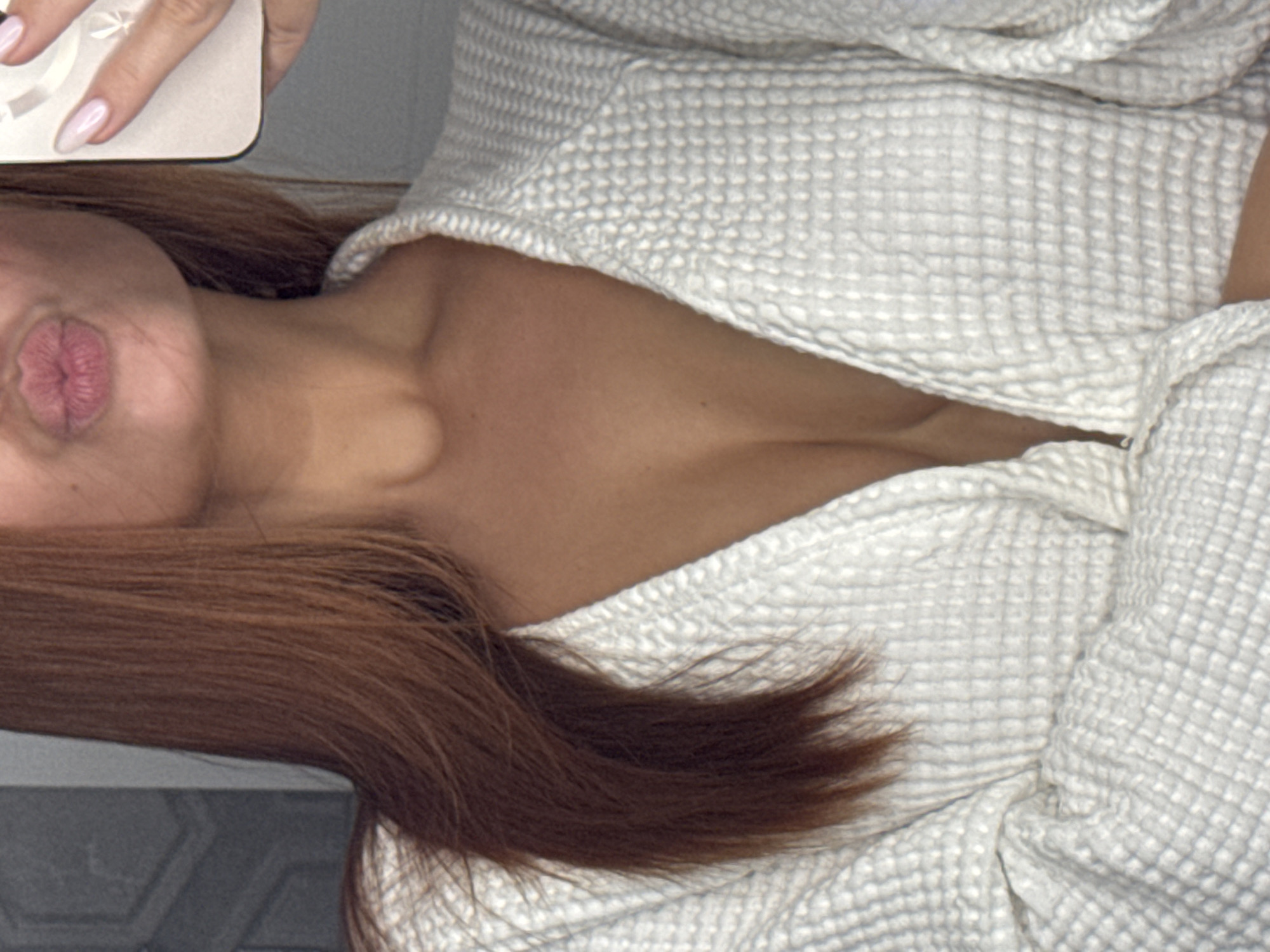 A person wearing a white waffle-weave robe is visible in the image. The person has auburn hair. The person's face is turned to the side with pursed lips. One of the person's hands holding a phone is visible in the upper left of the image. The location is not discernible from the image.
