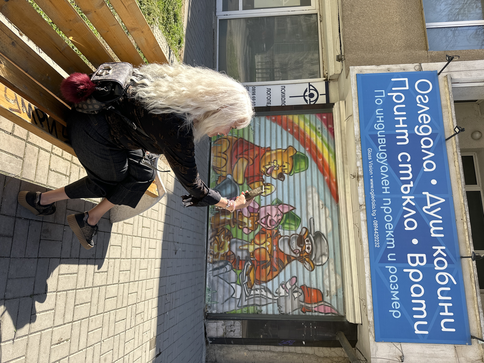 Here is a detailed description of the image:
An older woman with long, white hair is seated on a park bench next to a storefront. She is wearing a black shirt, dark pants, and dark shoes with platform soles. A dark purse with a red pom-pom is slung over her shoulder. She is holding a smartphone in her hands, seemingly taking a photo or interacting with the screen.
The storefront features a metal roll-down gate that is decorated with colorful cartoon characters, including Winnie the Pooh and others. Above the gate is a sign with writing in Cyrillic, as well as the words "психолог" and "логопед."
A large blue sign on the upper right side of the image, also with Cyrillic text, advertises services such as "огледала" (mirrors), "душ кабини" (shower cabins), "принт стъкла" (print glass), and "врати" (doors), specifying that they can be made "по индивидуален проект и размер" (by individual project and size). It also includes a website and phone number.
The scene is set on a paved sidewalk with sunlight casting shadows. Based on the Cyrillic text and architectural style, it is likely that the image was taken in a city in Bulgaria, potentially Sofia.