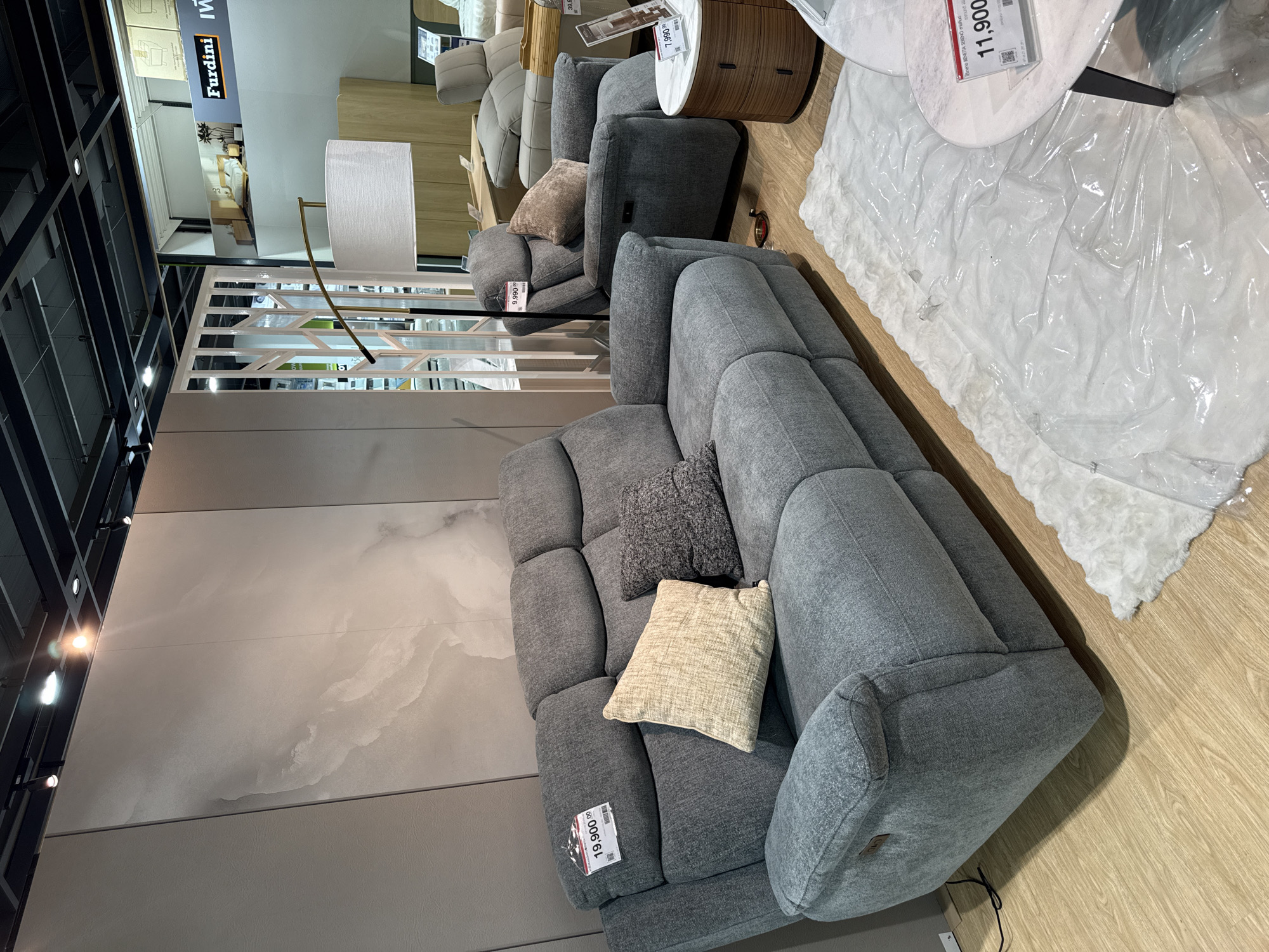 In the city of Baan Chaweng Noi, Thailand, a furniture showroom features a display of recliners and a sofa. The foreground is dominated by a gray fabric sofa with three headrests and two armrests. Two throw pillows, one light beige and textured, and the other dark gray and textured, are placed on the sofa. A price tag displaying "19,900.00" is attached to the left headrest of the sofa. To the right of the sofa is a single gray recliner with a small, light brown throw pillow on its seat. A price tag displaying "9,990.00" is attached to the headrest of the recliner.
In the background, a tall floor lamp with a white fabric shade and a metallic stand is positioned behind the sofa. Further back, a rounded wooden side table with two drawers is visible, topped with a round white marble coffee table. A price tag displaying "7,990.00" rests on the coffee table. A white shag rug, partially covered with clear plastic sheeting, is spread on the wooden floor in front of the furniture. The showroom walls are a neutral gray, with a large abstract art piece featuring marble-like patterns on the left. In the far background, a section of a display wall with the "Furdini" logo and an image of a bedroom scene is visible, along with other furniture items and shelving. The ceiling has exposed metal beams and track lighting. The scene appears to be indoors and well-lit, characteristic of a retail environment. No people are visible in the image.