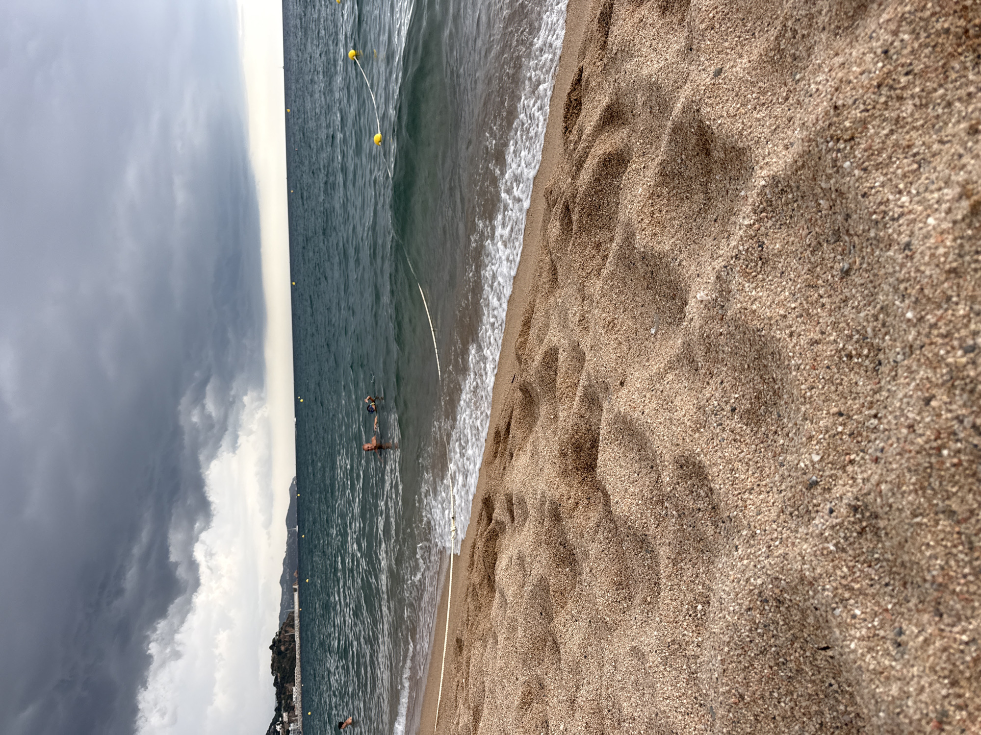 The image depicts a beach scene in Blanes, Spain, on a cloudy day with a dark, overcast sky dominating the upper portion. The foreground features a sandy beach with ripples and textures from the sand. The sea is visible, with waves gently lapping the shore and a white rope with yellow buoys marking a boundary in the water. In the middle ground, two individuals are in the water. One person is swimming or floating on their back, while another person appears to be partially submerged, possibly with a child or a companion. In the background, a coastline with buildings and vegetation can be seen, along with distant hills or mountains under the heavy clouds. The overall atmosphere is moody and potentially indicative of approaching rain.