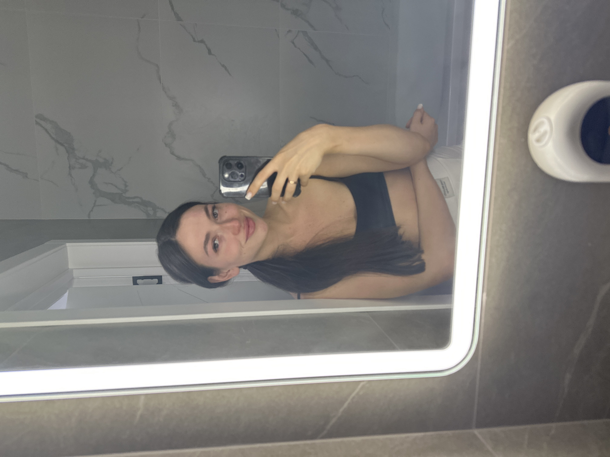 The image shows a young woman taking a mirror selfie in a bathroom. She has long, dark hair pulled back into a ponytail and is wearing a black sports bra and white shorts. She is holding a smartphone in her right hand, with her left hand resting on her thigh. The bathroom features modern grey marble-patterned tiles on the walls and a large, brightly lit mirror. The lighting is bright and even, suggesting an artificial light source. There is a white bathtub visible in the background, partially obscured by the reflection. The overall impression is of a clean and contemporary bathroom space. The location in the city of Эстачи, Russia is not discernible from the visual information present in the image.