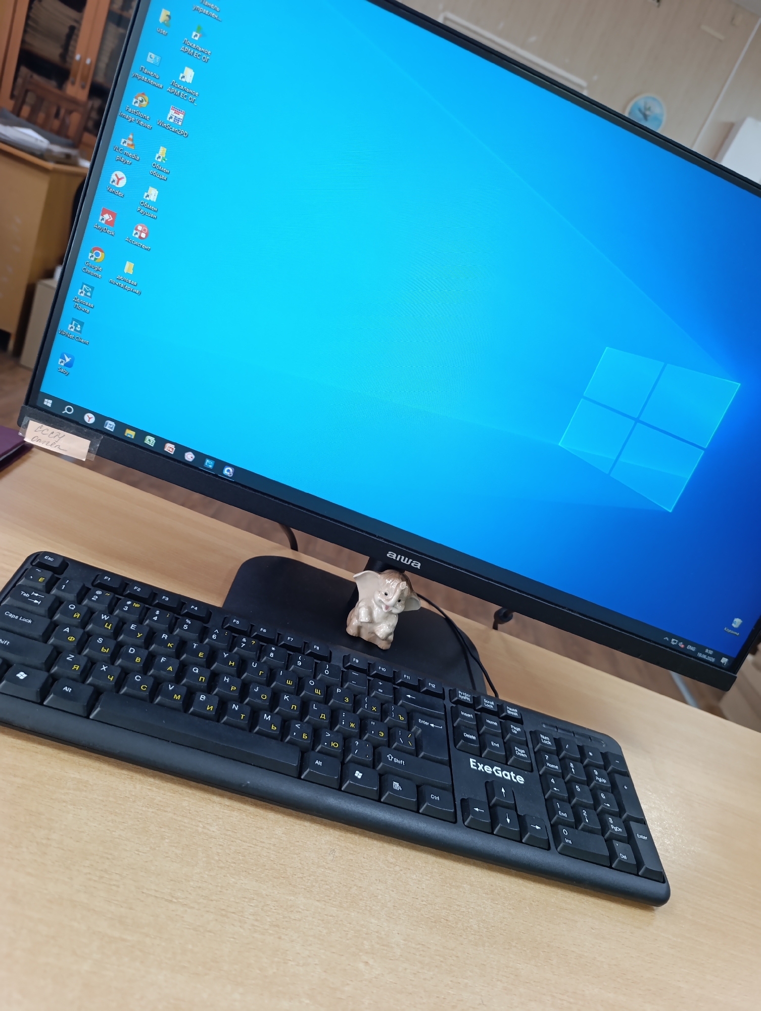 A computer monitor displays a vibrant blue Windows 10 background with the operating system's logo in the center. The monitor sits on a wooden desk, positioned in front of a black keyboard. A small, light-colored figurine, possibly a ceramic animal, is placed on the lower right corner of the monitor's bezel. The keyboard, labeled "ExeGate," features a standard QWERTY layout, with some keys showing Cyrillic characters. A piece of taped paper with handwritten text is affixed to the lower left bezel of the monitor. The background of the image reveals a blurred interior setting with bookshelves and office furniture, suggesting it's an office or study environment. The overall scene is static and captures a typical workspace.