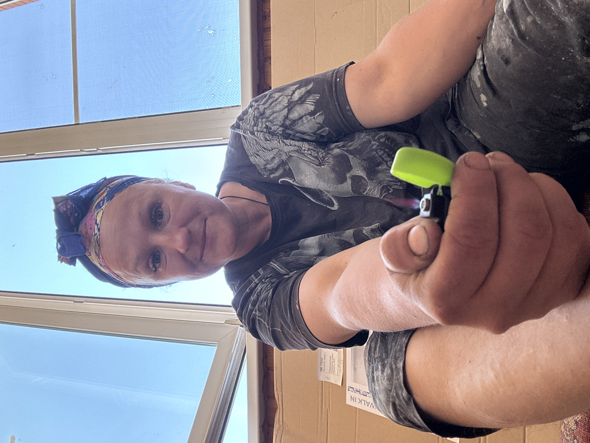 A woman is holding a lit lighter in her hand, viewed from a low angle. The lighter is black with a bright green cap. The woman has a colorful headband and is wearing a dark t-shirt with a skull design. Part of a window and sky are visible in the background. A cardboard box with text partially visible, including the word "WALK-IN", is behind the woman. The location is Chaikovsky, Russia.