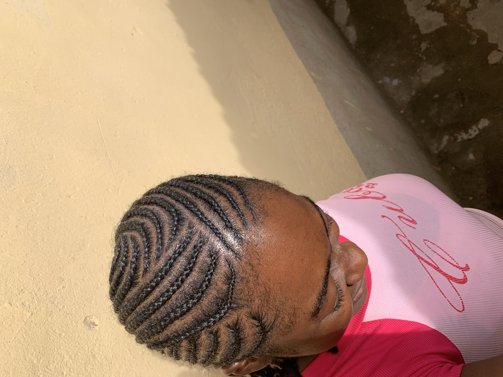A top-down view depicts a person with cornrow braids, their head slightly tilted towards the right. The braids are a dark color and arranged in a curved pattern, starting at the front of the head. The person is wearing a pink top, and part of their face is visible. The background consists of a light yellow wall and a dark, textured surface, possibly a concrete floor or wall. The location is Rafin Par, Nigeria.