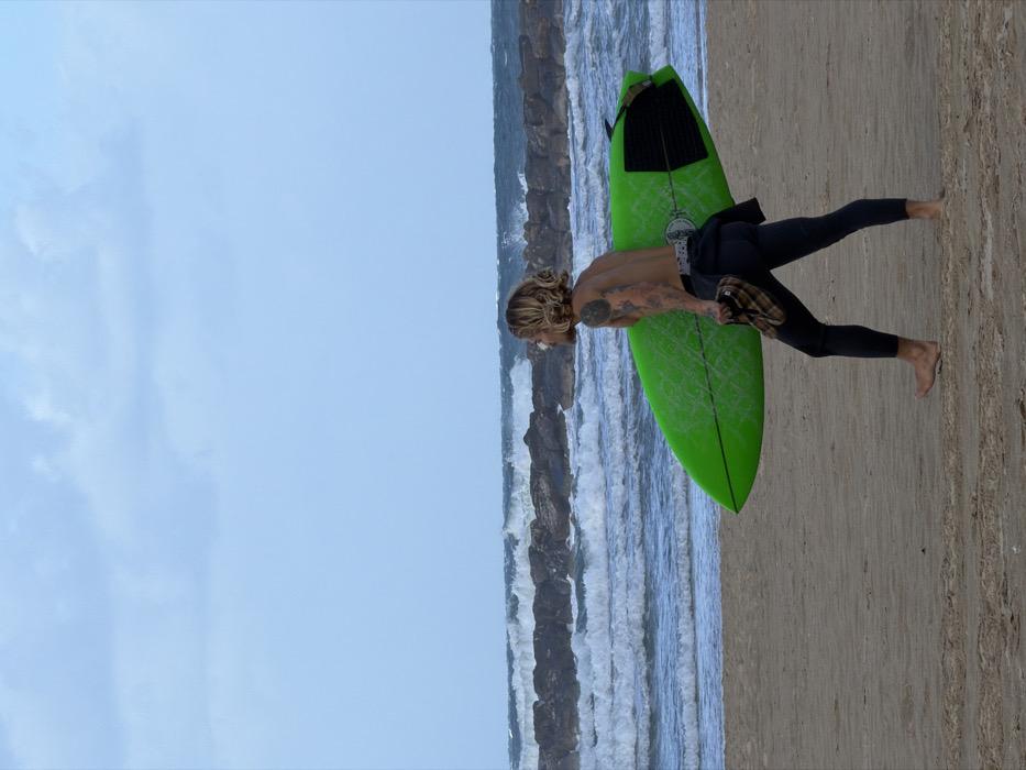 A shirtless surfer with tattoos carries a bright green surfboard on a beach. He wears a black wetsuit and holds what appears to be fins in his hand. The ocean with a rock barrier runs parallel to the sandy beach. A light blue sky fills the remaining space.