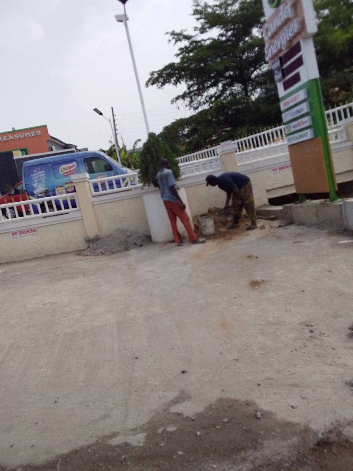 The image depicts an outdoor scene in what appears to be Lokoja, Nigeria, judging by the visible signage. Two men are working near a low white and tan wall with a "NO SMOKING" sign. One man wears a blue shirt and orange trousers and appears to be standing near a pile of gravel. The second man, wearing a dark shirt and patterned trousers, is bent over, seemingly mixing something in a bucket. Behind them is a tall sign that reads "Candle Energies," along with the words "WIN MART," "Car Wash," "NTS," and "RESTAURANT." A blue van with "Checkers" branding is parked behind the wall. The ground is concrete. A tall streetlight and trees are visible in the background. The sky is overcast.