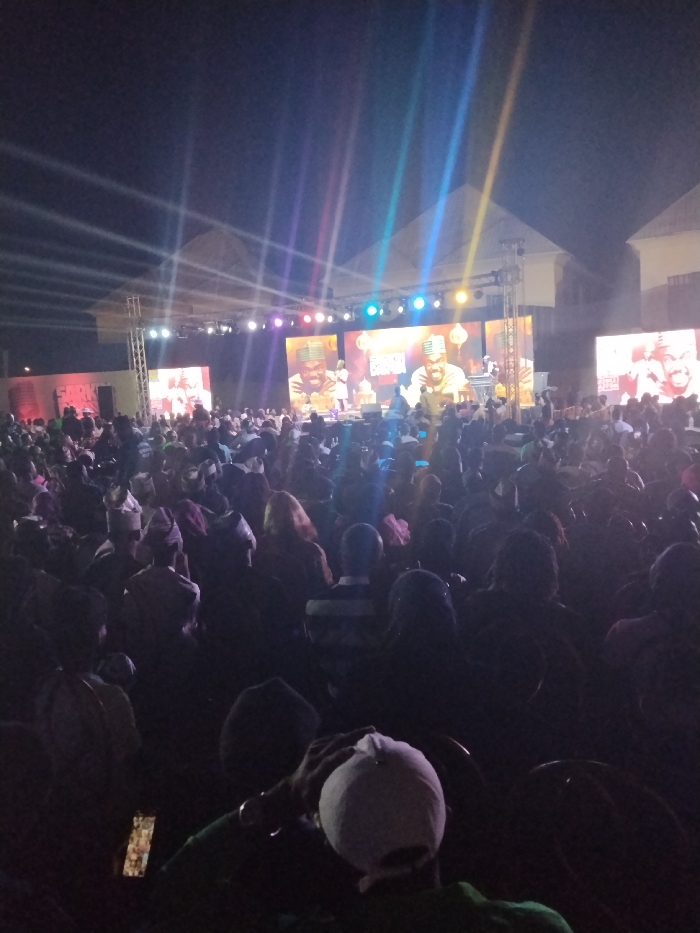 The image depicts an outdoor nighttime event taking place in Fagge D2, Nigeria. A large audience, consisting of hundreds of individuals, is gathered in an open area, predominantly seated in rows of chairs, facing a brightly lit stage structure.
The stage features a metal truss framework supporting multiple large LED screens. The central screens display a consistent image of a bearded male figure wearing a cap or head covering, holding a microphone, suggesting a performance or speech. Stage lighting fixtures are mounted on the truss, emitting visible beams of colored light (blue, green, purple, yellow, white) that extend upwards into the dark night sky. Additional screens are visible to the left and right of the main stage.
In the foreground, the backs of audience members are visible, mostly silhouetted against the stage lights. One individual in the immediate foreground wears a white baseball cap. Another person holds an illuminated mobile phone. Behind the stage, parts of buildings with light-colored roofs are discernable. The crowd extends into the deep background, indicating a substantial attendance.