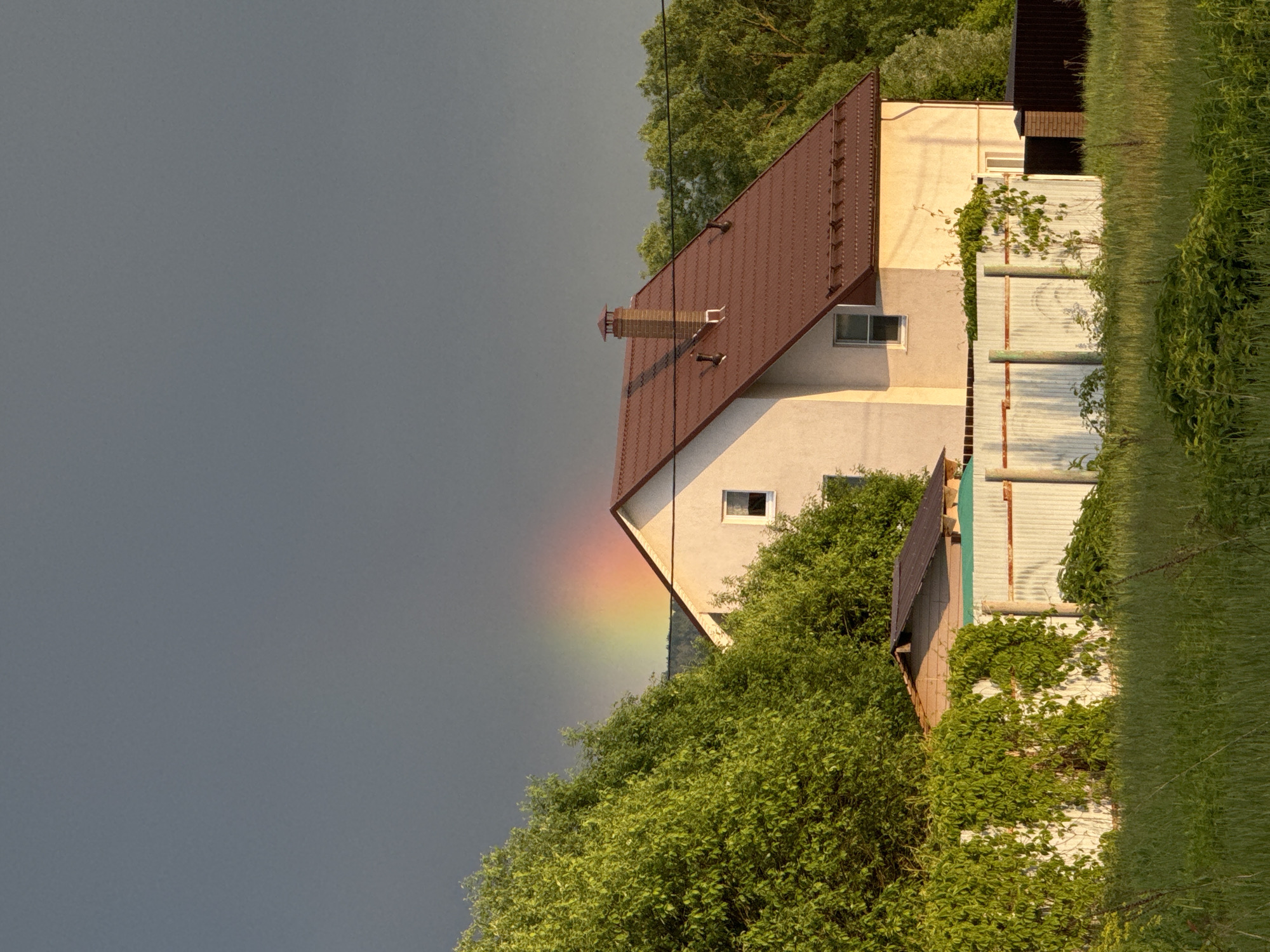The image shows a house in Vysokinichi, Russia, with a partial rainbow visible on the left side of the building against a dark, overcast sky. The house is two stories with a brown roof, a chimney, and white siding. A white fence with vines growing on it runs along the right side of the house. Lush green foliage surrounds the house, partially obscuring it from view. A small outbuilding with a dark roof is situated next to the house. The foreground includes grass and more dense foliage.