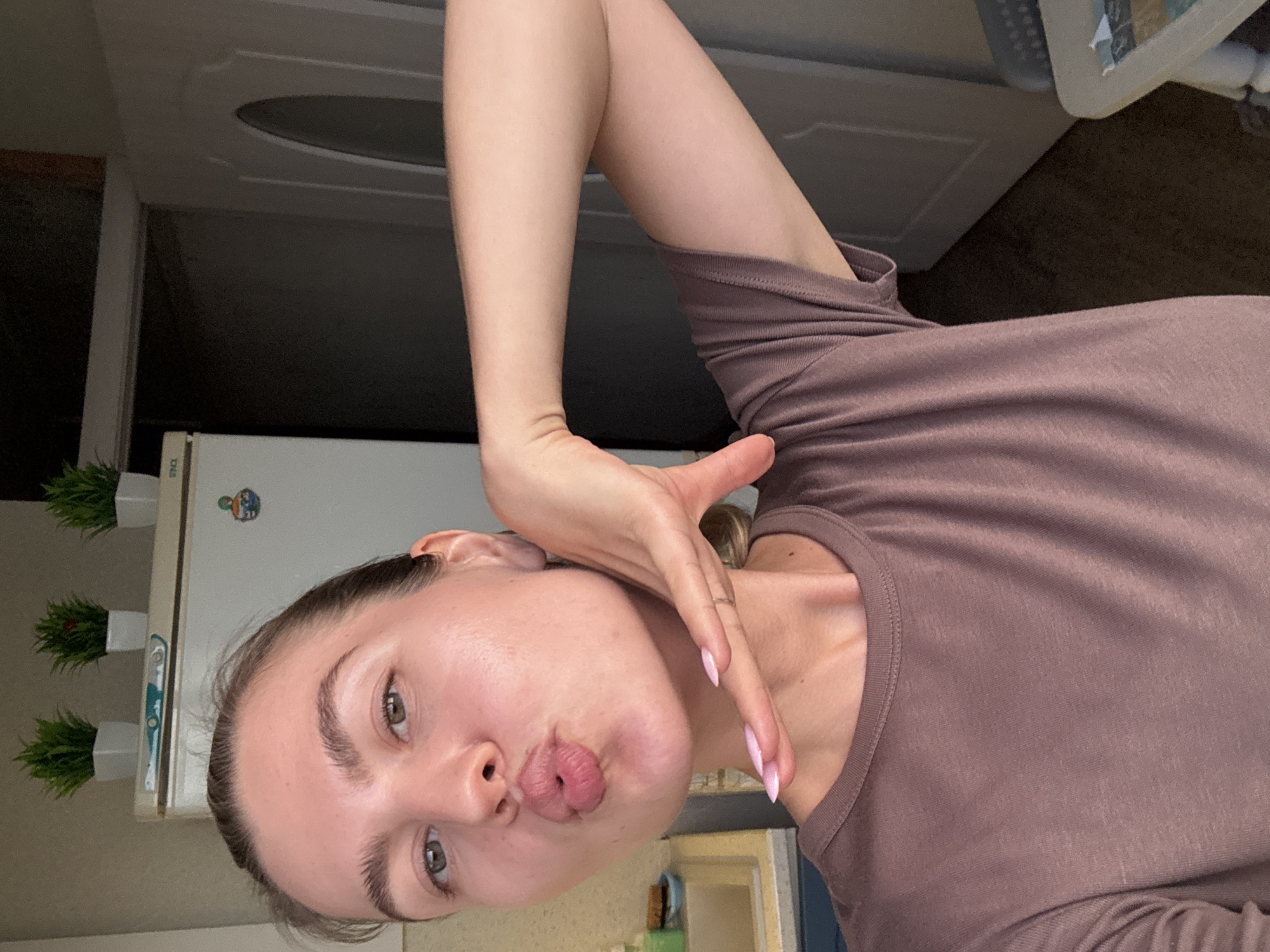 An indoor, close-up shot features a young woman with light skin and dark brown hair pulled back from her face. She is looking directly at the camera and puckering her lips in a kiss. Her right arm is bent, with her hand raised to her cheek, and her fingers are adorned with light pink nail polish and a silver ring. She is wearing a brown t-shirt. In the background, a white refrigerator is visible with three small potted plants on top. To the right of the refrigerator, a dark doorway and a portion of a white door can be seen.