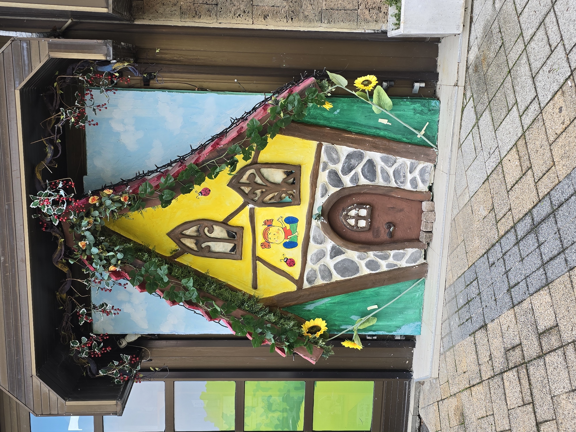 This image captures a vibrant, whimsical, miniature house facade displayed outdoors. The house is brightly colored, featuring a dominant yellow section with two arched, ornate brown-framed windows in its upper half. Below one window, a cartoon drawing of a child in blue overalls and a ladybug adds to the playful theme. The right side of the house is adorned with a gray stone pattern, leading to a small, arched brown door with a tiny window. The base of the house is painted a bright green.
The roofline of the house is decorated with artificial green ivy, small red berries, and strings of tiny lights, suggesting a festive or enchanted atmosphere. Additional vines with red berries cascade down the left side of the display. Several artificial sunflowers on green stems protrude from the right side and near the bottom, enhancing the natural, cheerful feel. The immediate background behind the house is a painted blue sky with soft white clouds.
The entire decorative piece is set against a larger building structure. Brown wooden panels are visible above and to the left of the house, while windows with green-tinted glass are present below. To the right, a paved sidewalk with rectangular blocks of alternating grey and beige provides a clear pedestrian path. The scene appears to be well-lit, indicating it is daytime. The installation creates a charming, storybook-like impression, likely serving as an artistic display or a unique shop front. No discernible text is visible within the image.
