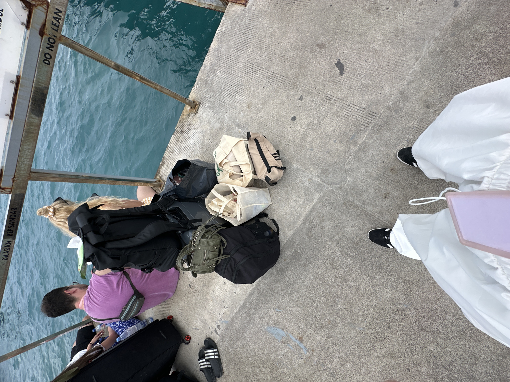 The scene is on a concrete pier in Kampung Toyapakeh, Indonesia, beside a body of water. There are several pieces of luggage and bags, including black backpacks, canvas tote bags, and a rolling suitcase. Two people are visible: one wearing a purple shirt and the other wearing white pants and black shoes. A sign with the phrase "DO NOT LEAN" can be seen. The environment suggests a travel or waiting situation.