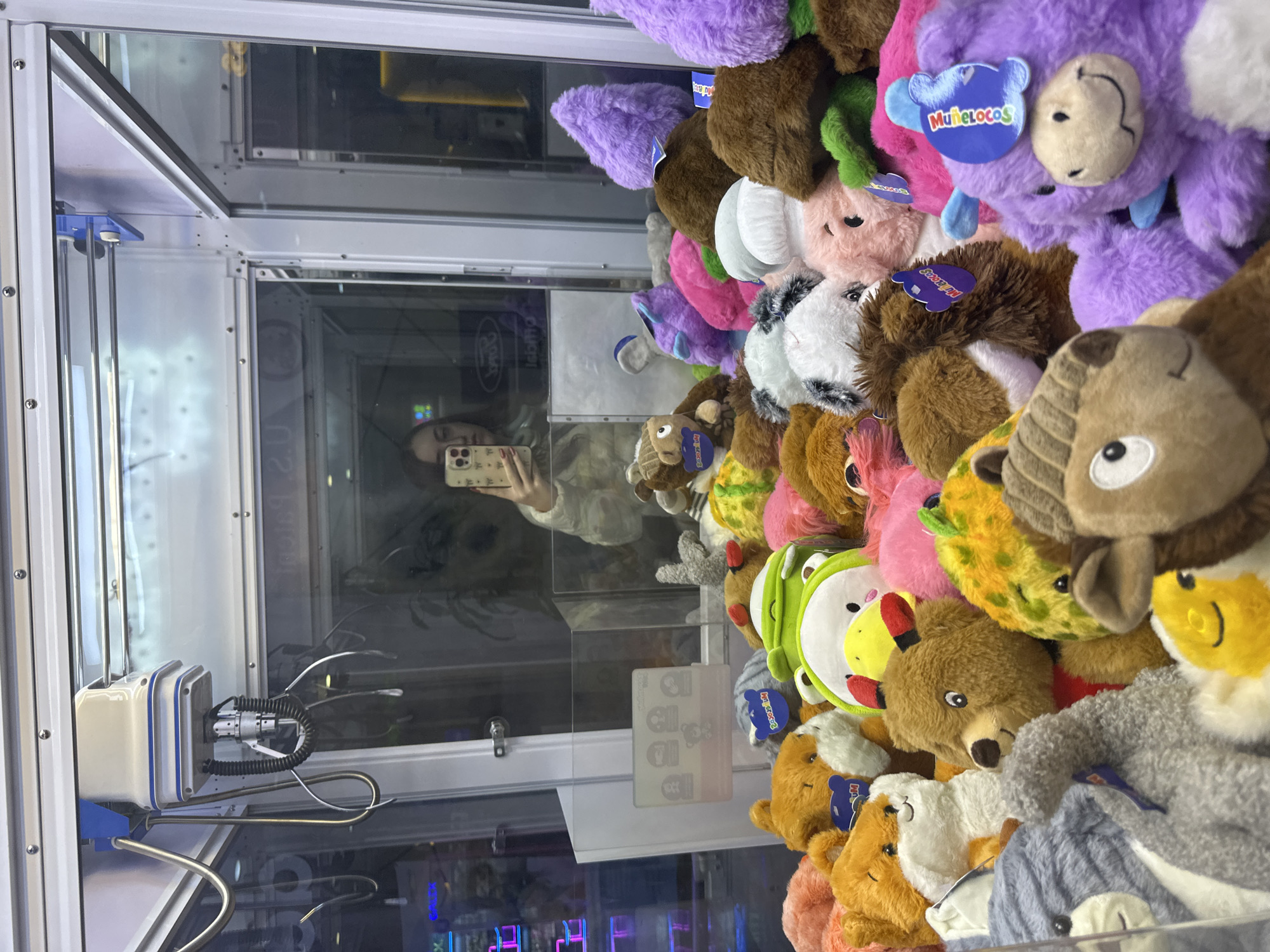 A view inside a claw crane arcade game filled with stuffed animals, reflected in the glass is a person holding a smartphone. The game is likely located in Huixquilucan de Degollado, Mexico, based on the tags on the stuffed animals which read "Muñelocos." The claw mechanism is visible on the left side of the frame.