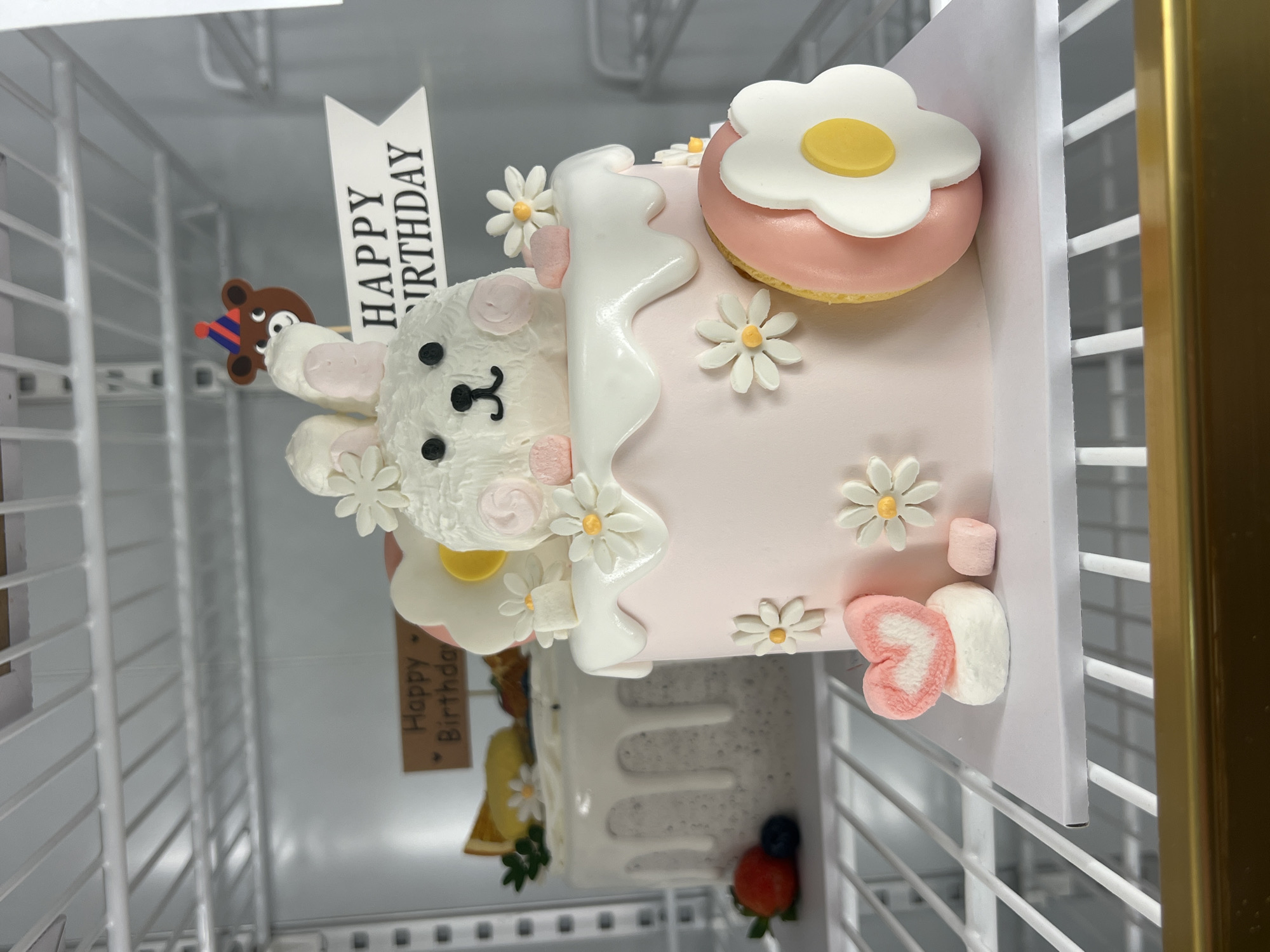A decorated birthday cake rests on a white surface inside a refrigerator. The cake is adorned with a white bear figurine, daisy-shaped candies, and a donut-shaped topping with a white flower and yellow center. White icing cascades down the side of the cake. A "Happy Birthday" sign is attached to the cake. The refrigerator has a white wire rack interior, and a gold frame is visible on the right side. Another cake with different decoration appears in the background. The photo was taken indoors. The city of Nha Trang, Vietnam is not deducible from the given information.