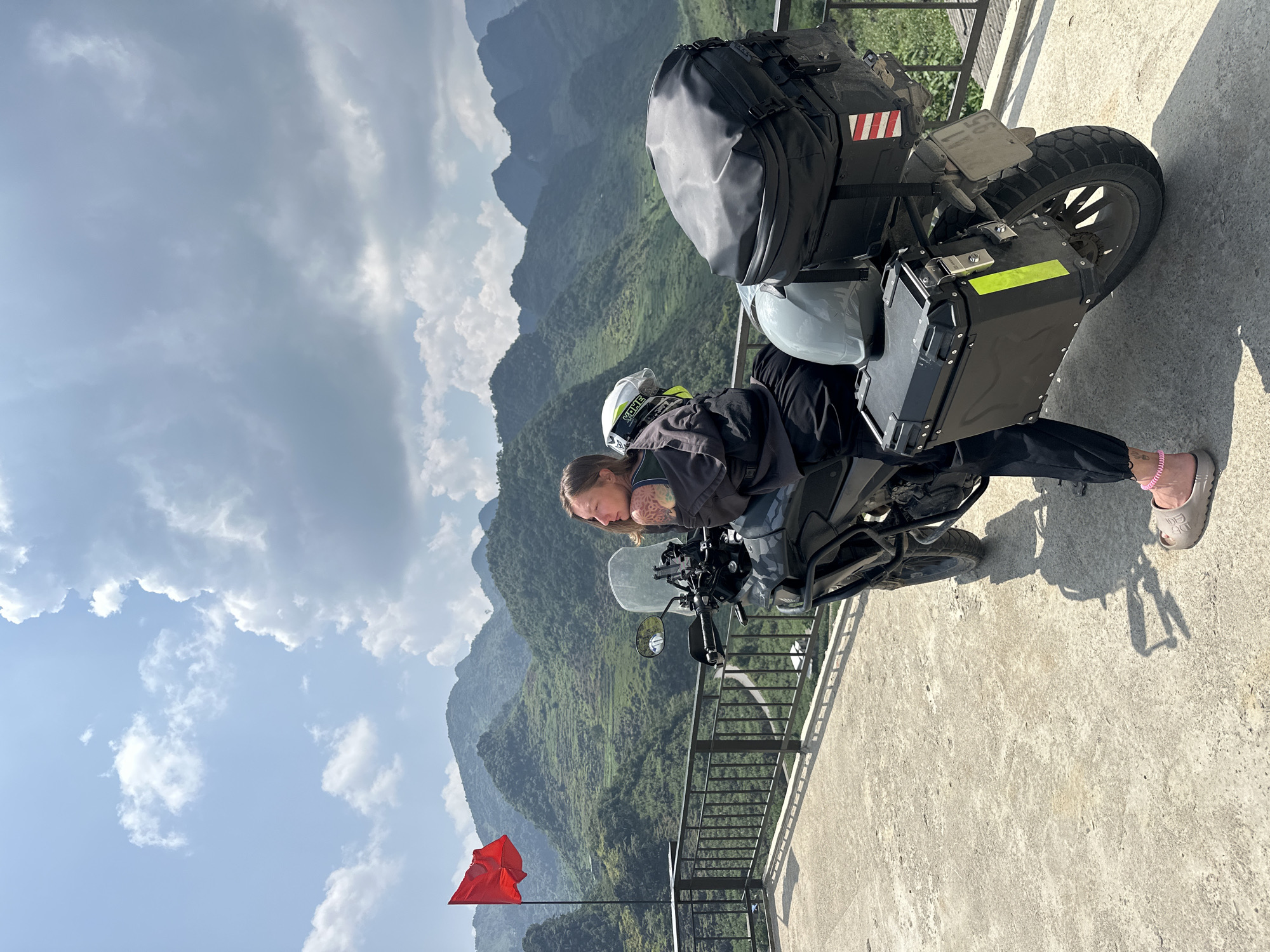 A person is sitting on a parked motorcycle on a concrete platform, with mountains and a partly cloudy sky in the background. The motorcycle is black with panniers, a top case, and a helmet strapped to the back. The person is wearing riding gear, including a jacket and a helmet. A red flag is visible in the lower left corner of the image. The location is Lũng Phìn Commune, Vietnam.